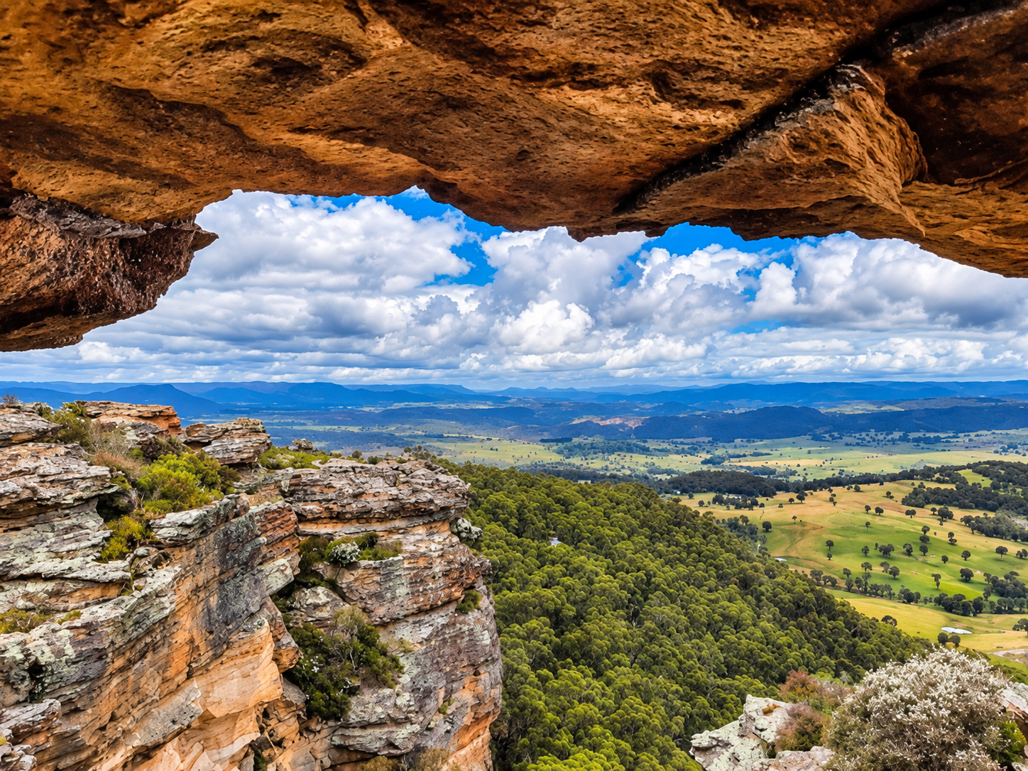 View of a landscape seen through a rocky overhang, showing green forested hills, fields, and a distant mountain range under a partly cloudy sky.