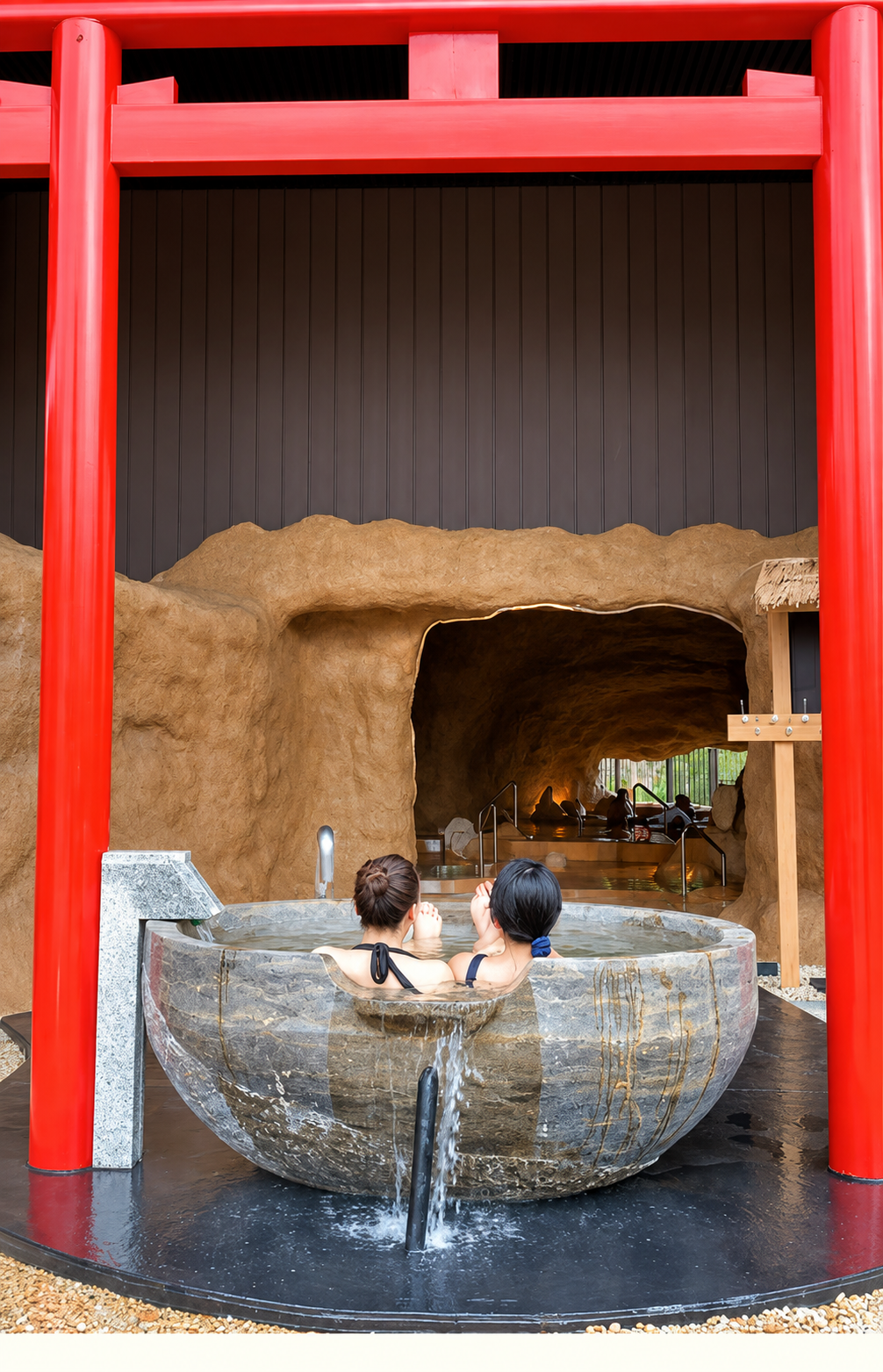 Two women soaking in a large, round stone hot tub with a cave-like interior and a black and red exterior structure.