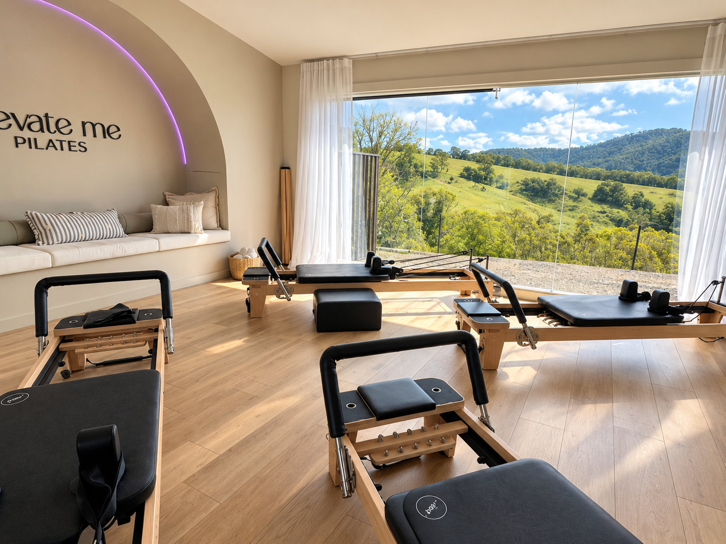 Pilates studio with reformer machines facing large window showing green hills and blue sky.