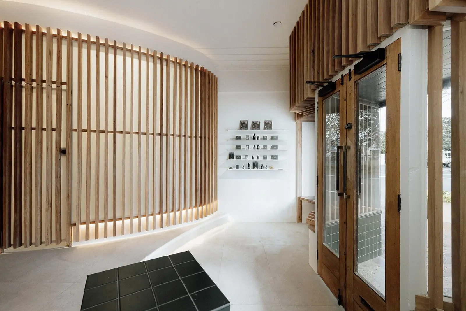 Interior of a modern space with wooden slat wall design, white shelves with small bottles, and a glass door with a wooden frame.