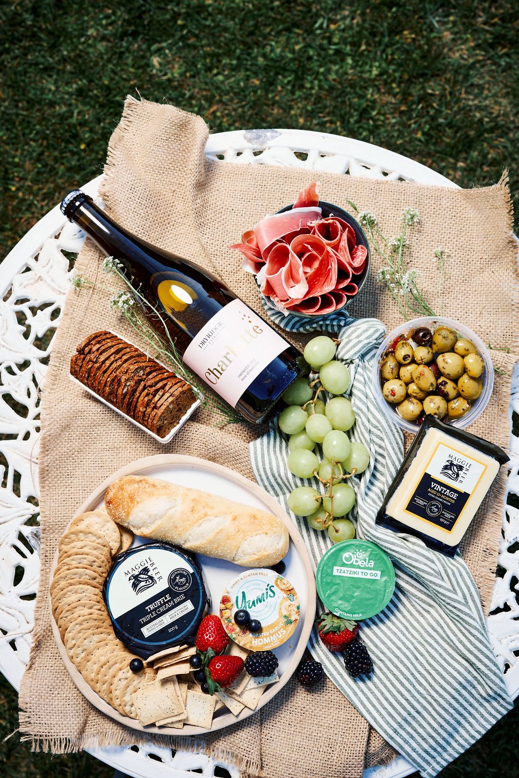 A picnic spread on a white metal table with a burlap cloth, featuring a bottle of dry sparkling wine, sliced cured meat, green grapes, a cheese wheel, assorted cheeses and crackers, mixed berries, green olives, black olives, and a container of hummus, with some small white flowers and strawberries.