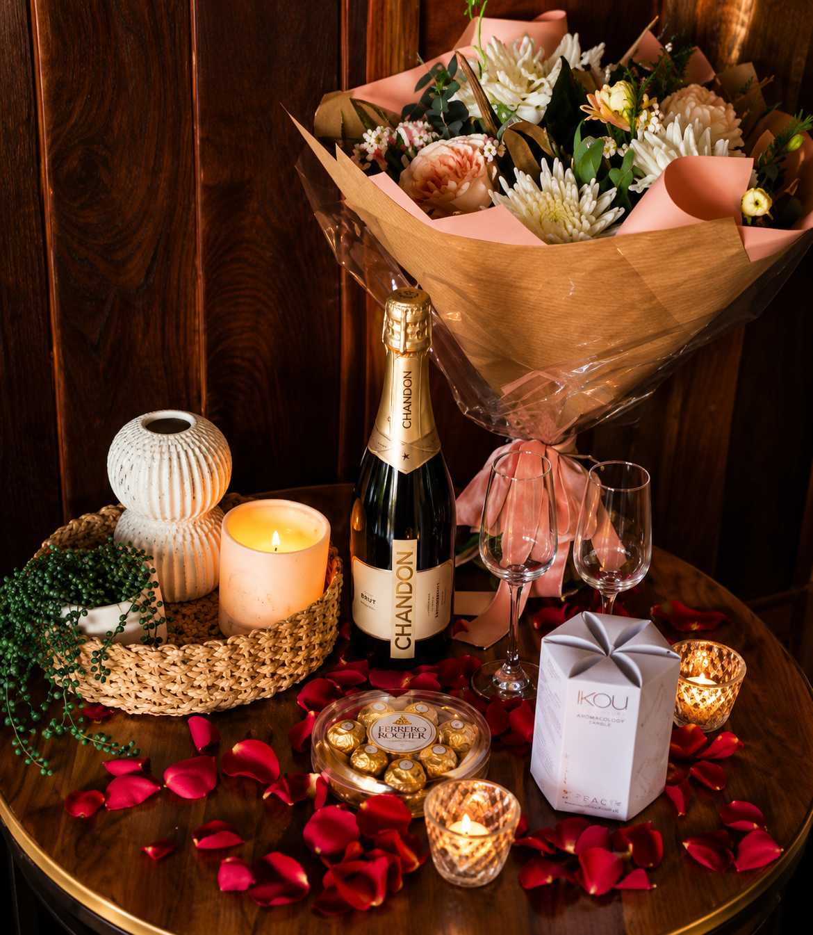 A romantic table setting with a bouquet of flowers, a bottle of Chandon champagne, two empty glasses, candles, flowers petals, chocolates, and decorative items on a wooden table.