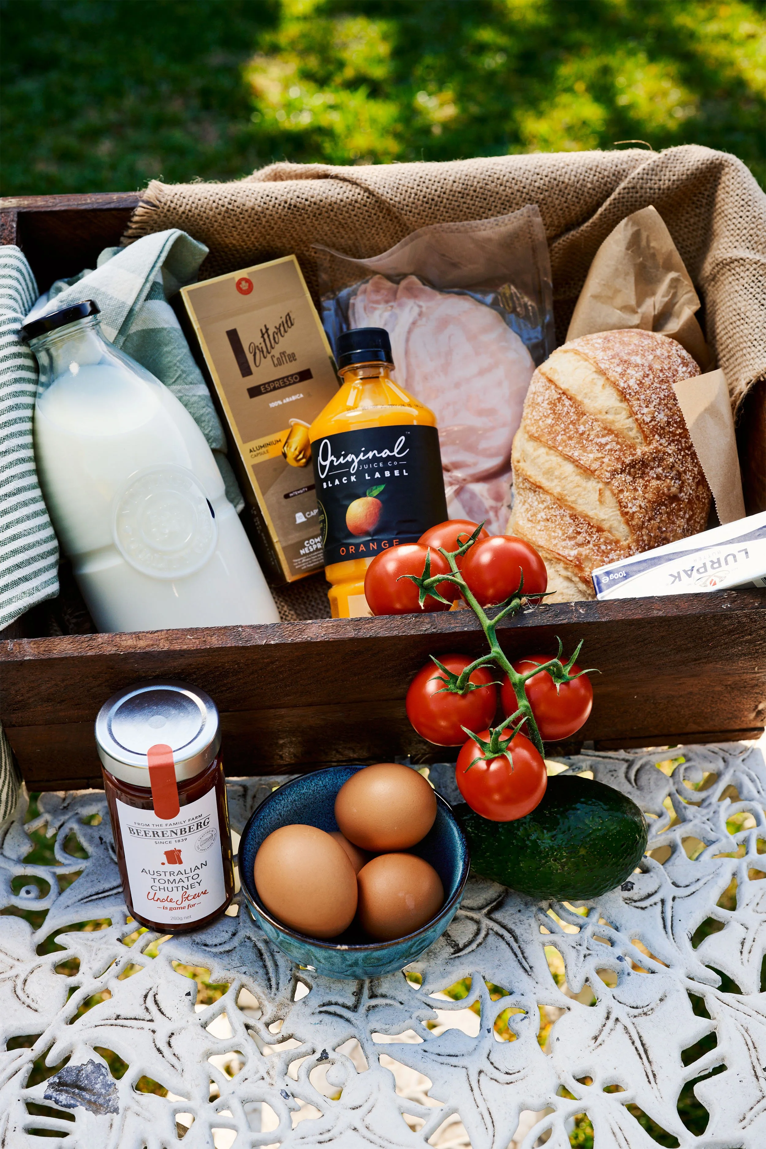 A picnic basket containing a jar of milk, a carton of coffee, a packaged piece of meat, a loaf of bread, cherry tomatoes, eggs, a cucumber, a jar of chutney, and an avocado, all arranged on a decorative metal outdoor table with green grass in the background.