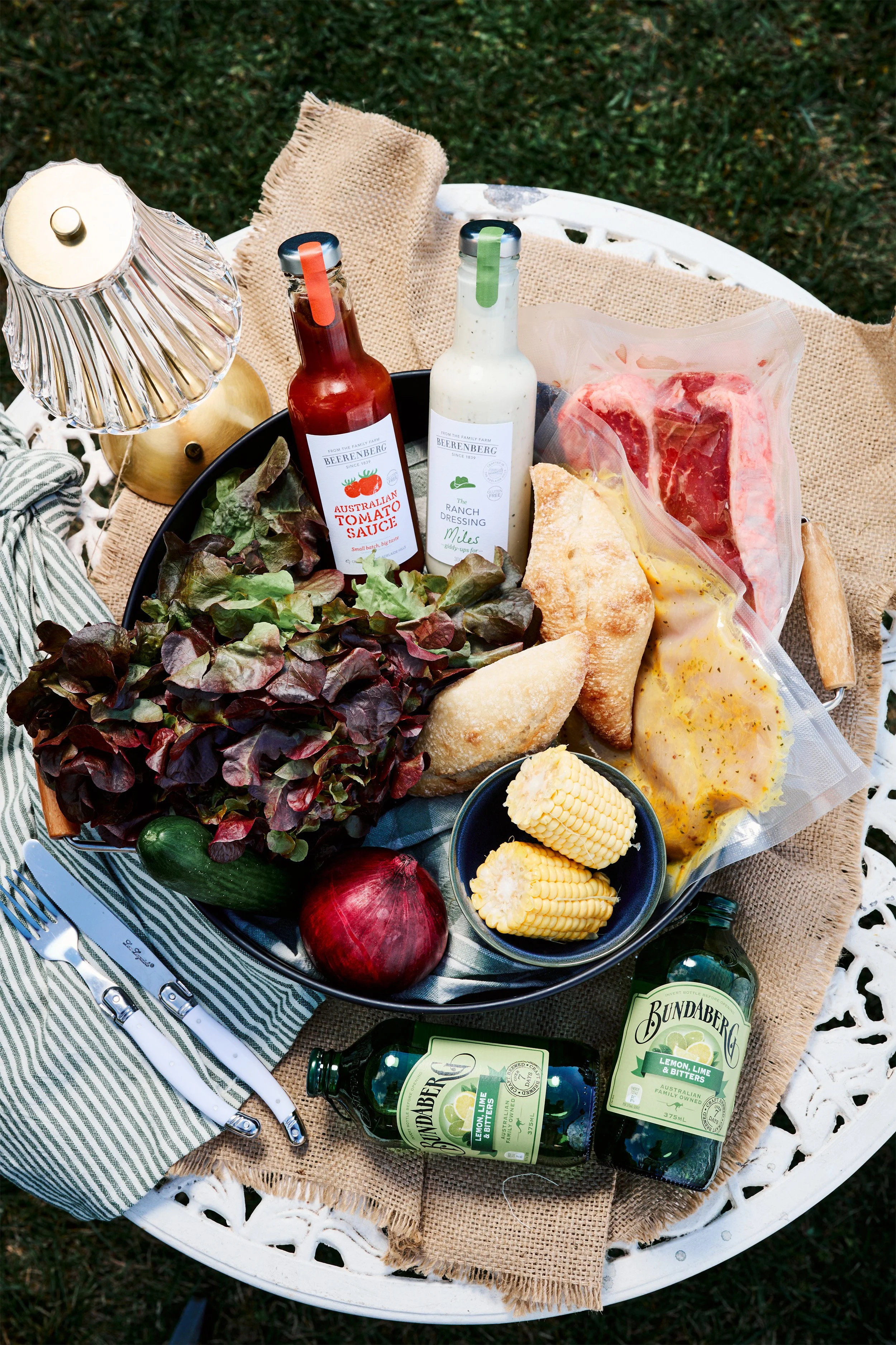 A picnic table setup with a black tray of fresh salad greens, cherry tomatoes, and corn, along with raw meat, bread, and condiments including tomato sauce and ranch dressing. There are two bottles of Bundaberg lemon lime and bitters, a red onion, a cucumber, a red apple, and an apple cut in half. A striped napkin with a fork and knife is also present, and the table is adorned with a burlap cloth, all outdoors on grass.