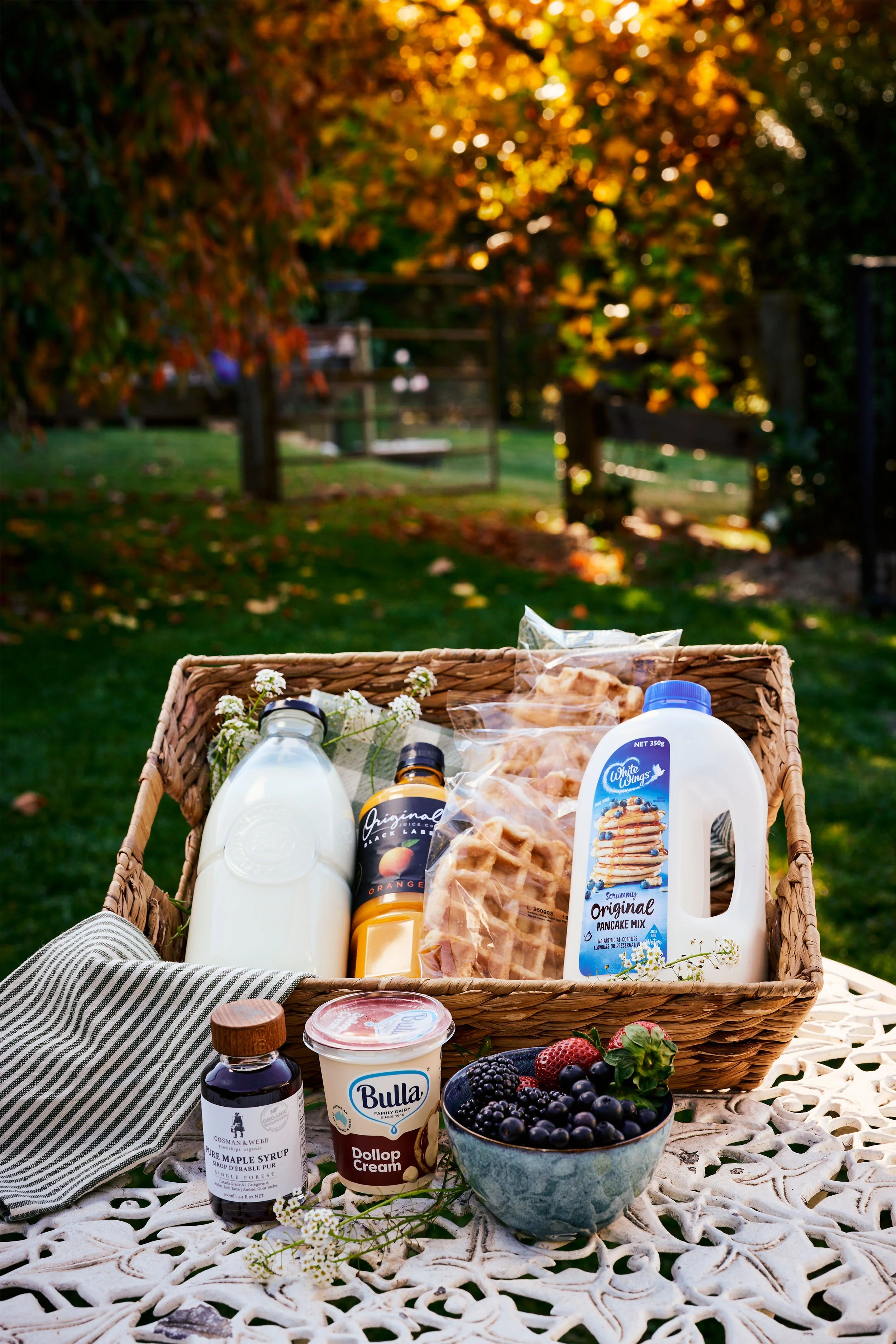 A picnic basket filled with bottles of milk, orange juice, pancake mix, and pancake waffles, with a bowl of mixed berries, a jar of syrup, yogurt, and small flowers on a lace tablecloth outdoors in a garden with trees and a fence.