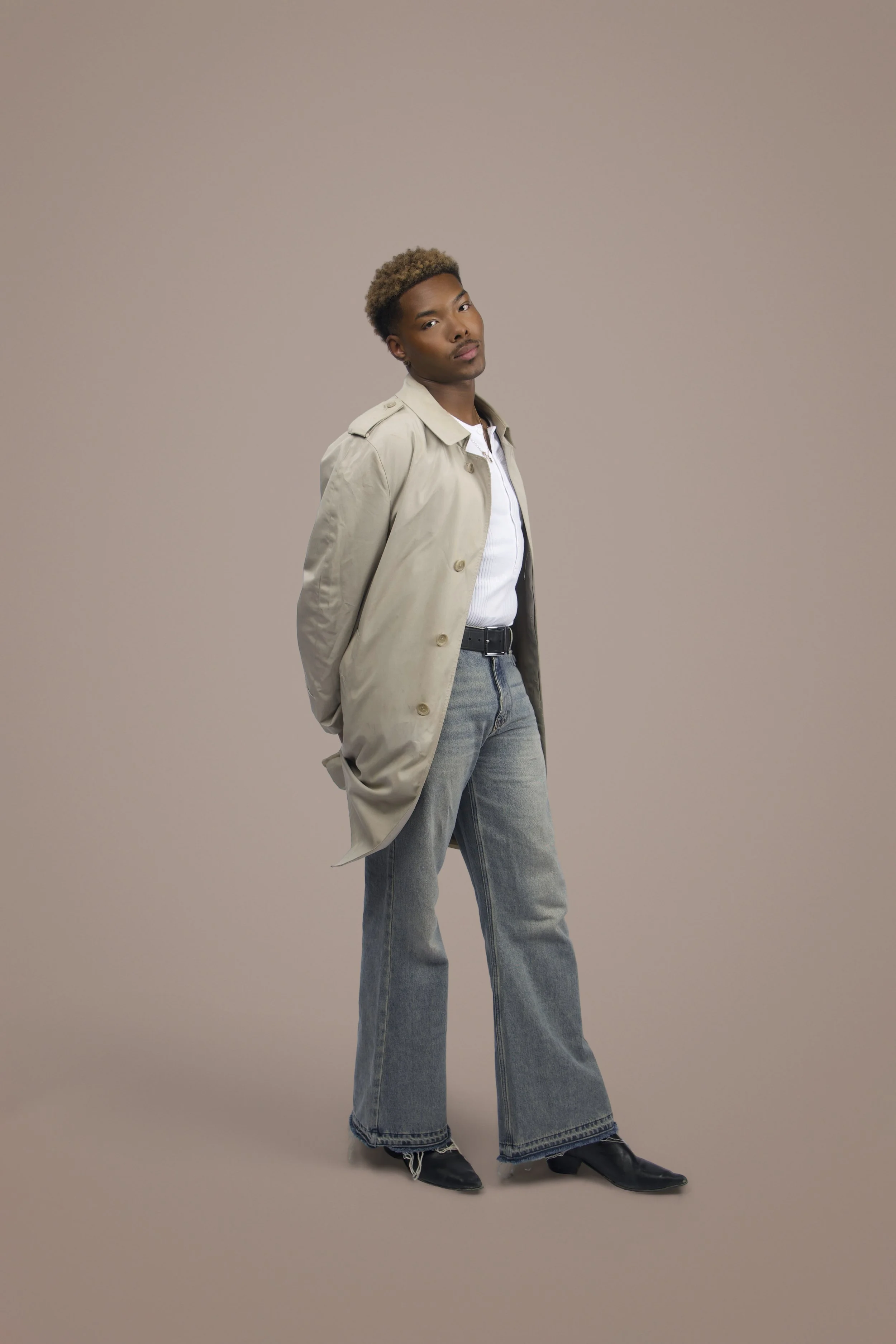 A young man with light brown skin and curly hair standing against a plain background. He is wearing a beige trench coat, a white button-up shirt, and high-waisted, wide-leg jeans. He has black pointed shoes and is looking directly at the camera with a confident expression.