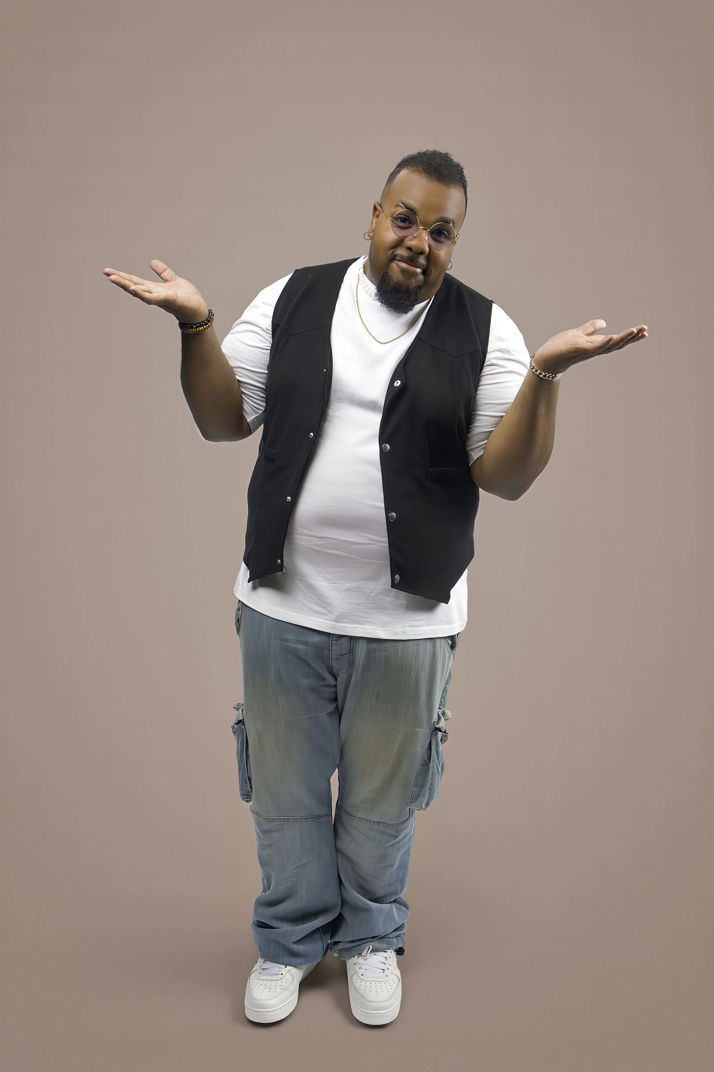 A person with short hair, beard, and glasses, standing with arms raised in a shrug gesture, wearing a white t-shirt, black vest, gray cargo pants, and white sneakers, against a neutral gray background.