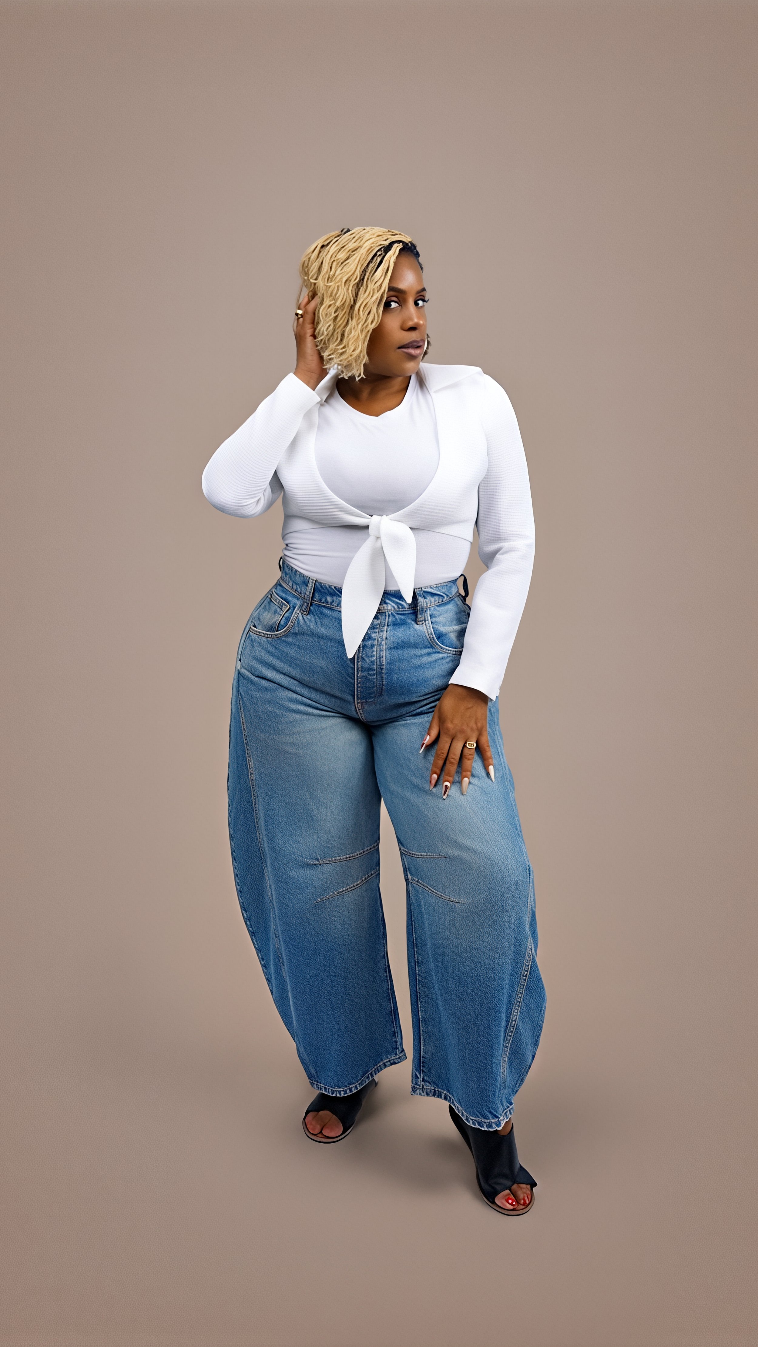 A woman with blonde muscle braids hairstyle wearing a white long-sleeve top with a tie front, high-waisted blue jeans, and black open-toe heels posing against a plain background.
