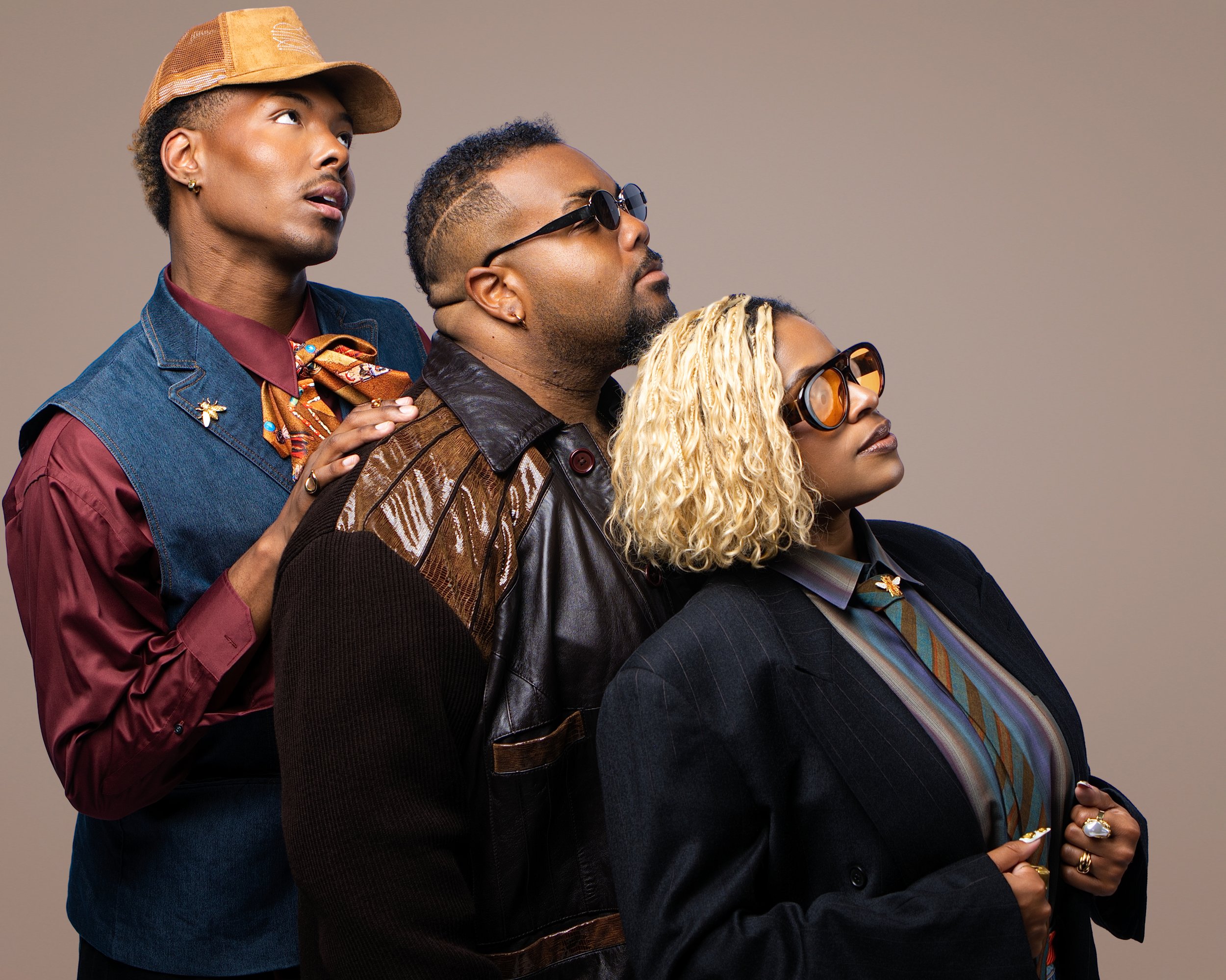 Group of four diverse individuals standing close together, looking upward. The person on the left wears a tan cap, denim jacket, and patterned scarf; the second wears sunglasses and a brown leather jacket; the third has blonde curly hair and sunglasses, and the fourth wears a dark blazer with a colorful striped shirt, all with confident expressions.