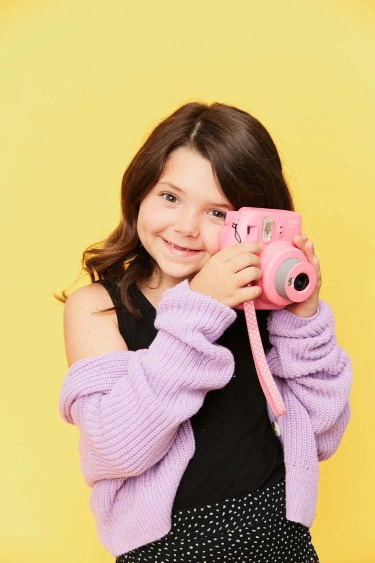 Houston School Photography | Young girl smiling and holding a pink instant camera against a yellow background.