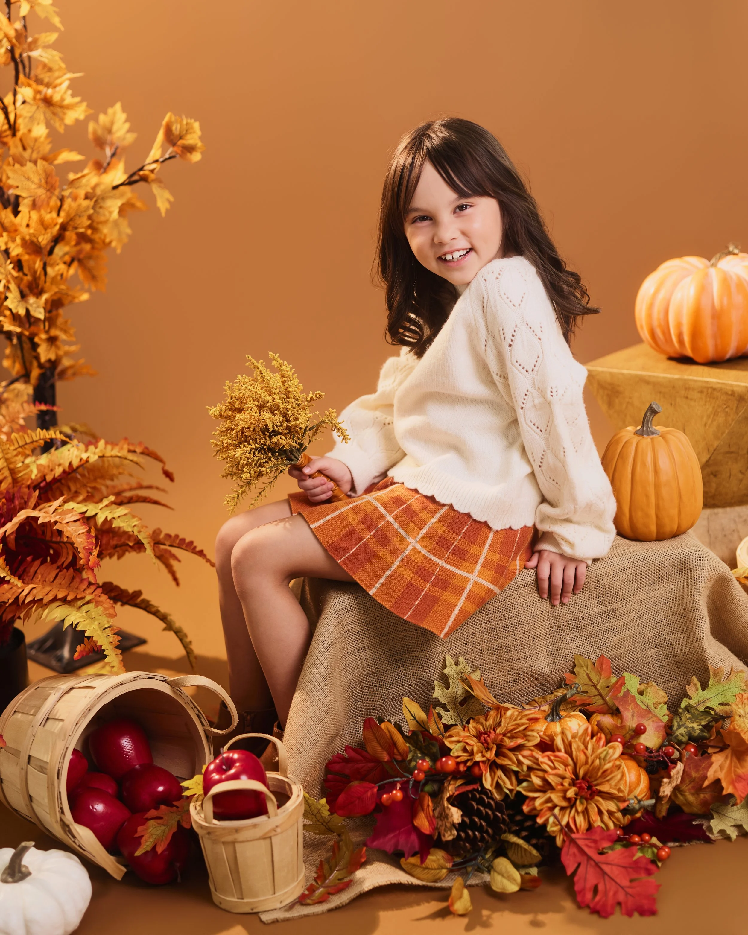 Houston Fall Mini Session | A young girl with dark hair and a white sweater smiling while sitting among fall-themed decorations including pumpkins, apples, autumn leaves, and dried flowers with a warm brown background.