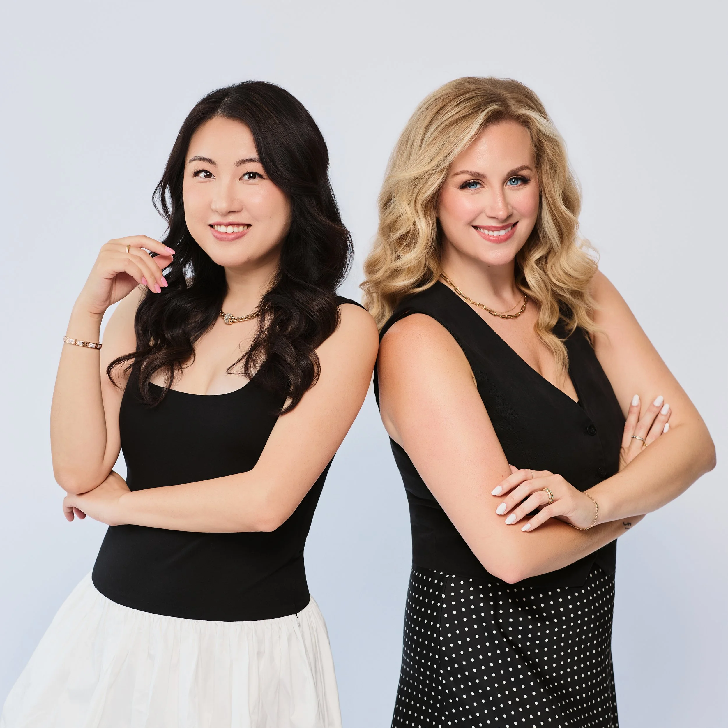 Best Houston School Photographer | Two women standing side by side with crossed arms, wearing black tops, one has a white pleated skirt and the other has a black and white polka dot skirt, smiling against a light background.