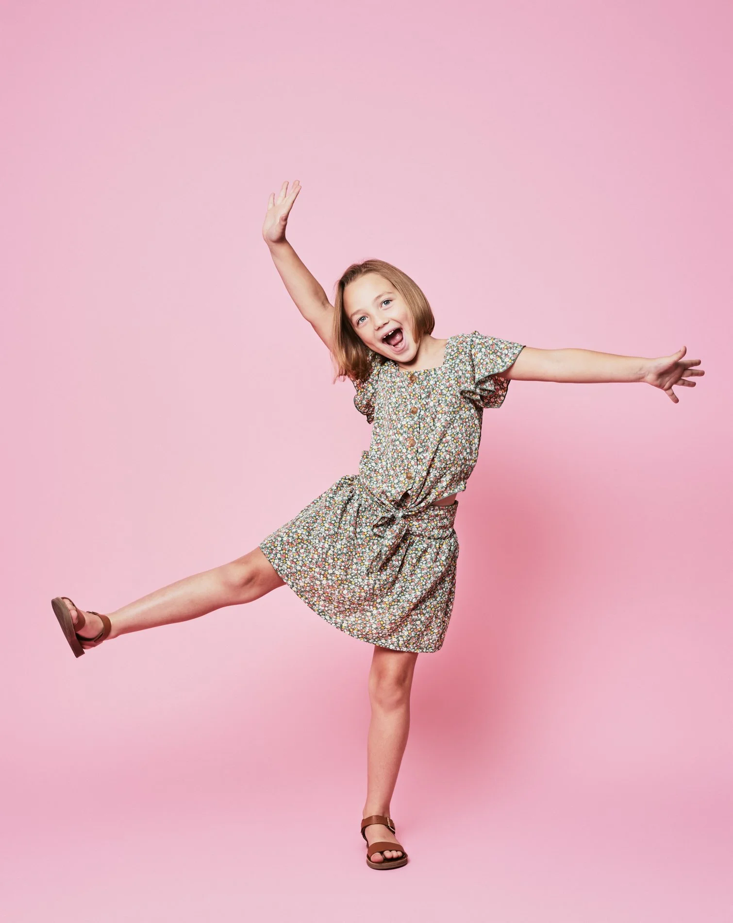 A young girl with red hair, wearing a floral dress, is jumping with one leg in the air and arms outstretched against a pink background.