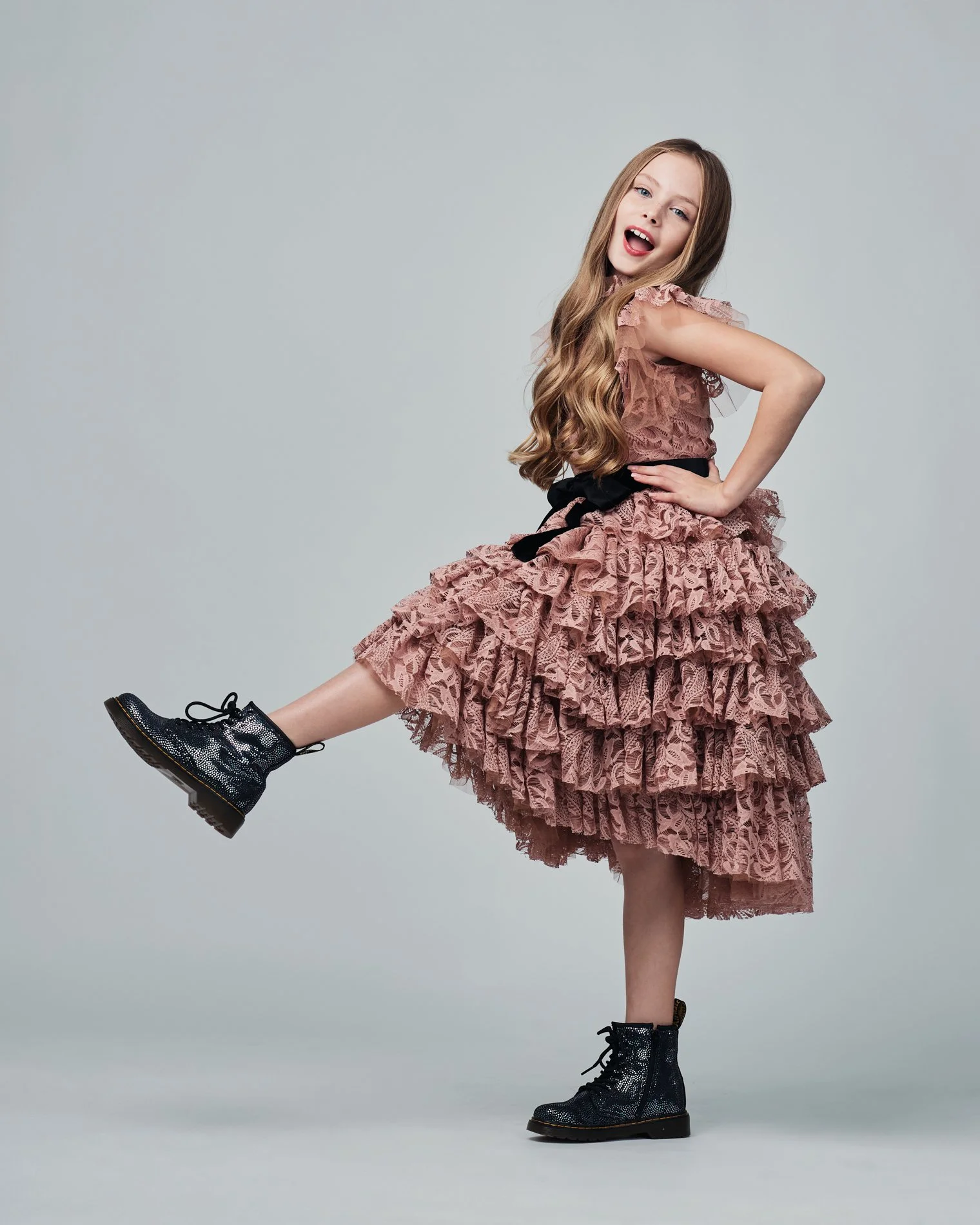 Young girl with long wavy hair, wearing a pink ruffled lace dress with black belt, black shiny lace-up boots, posing with one leg raised and hand on her hip, smiling against a plain light grey background.