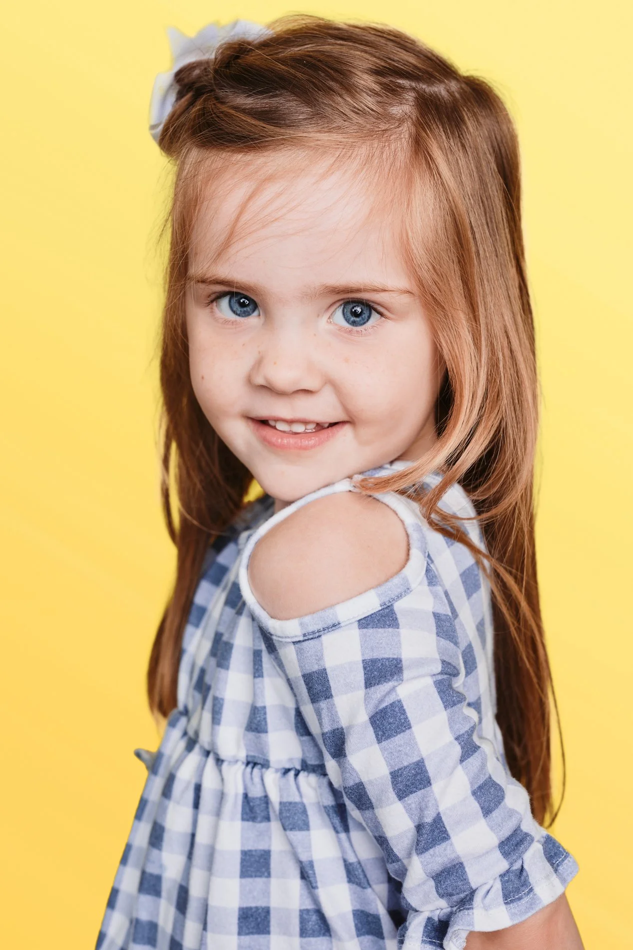 A young girl with red hair and blue eyes smiling, wearing a blue and white checked dress with cut-out shoulders, set against a yellow background.