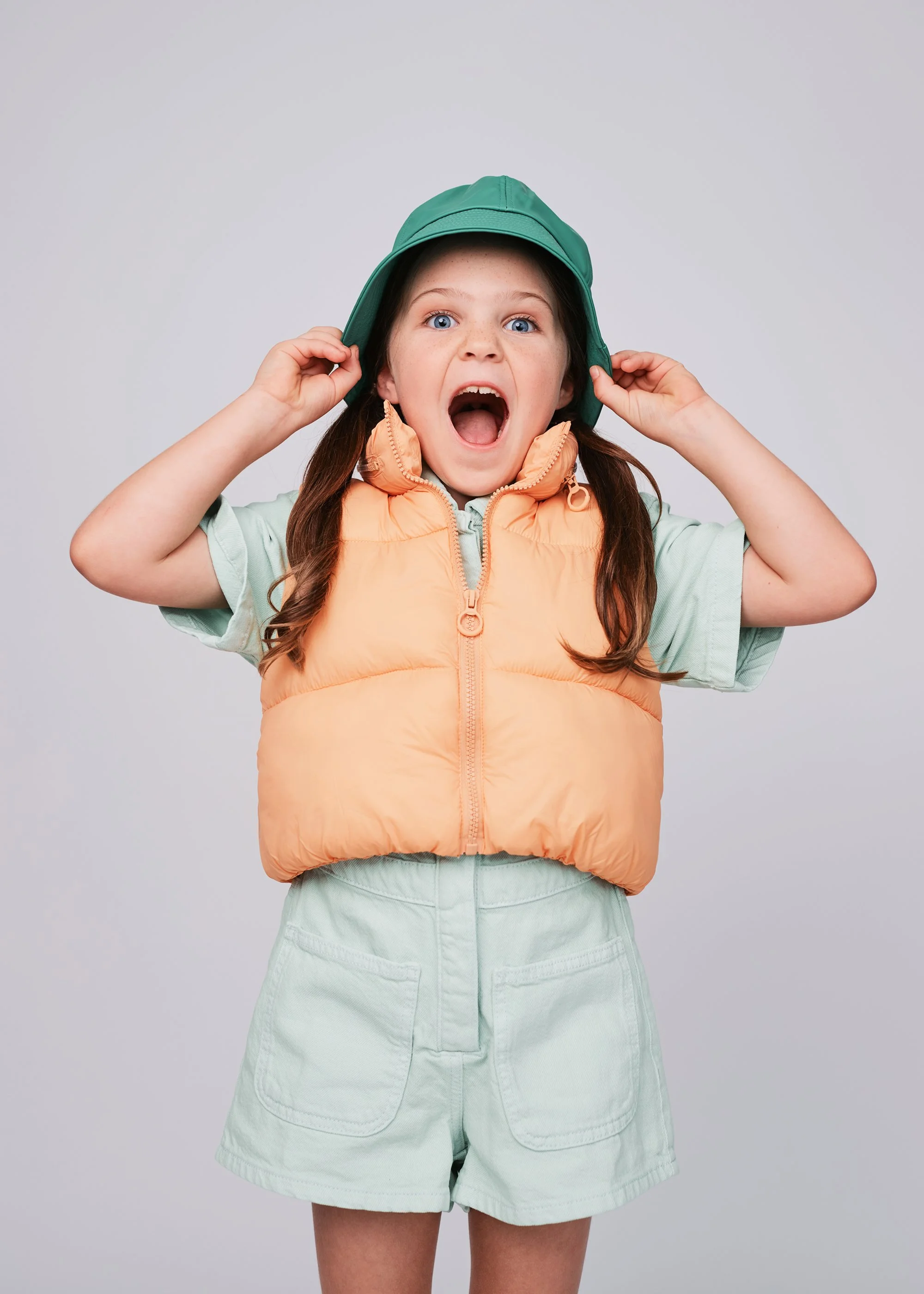 Young girl with blue eyes and brown hair, wearing a green hat, a light green shirt, a peach puffer vest, and light green shorts, posing with mouth open and hands on her hat.