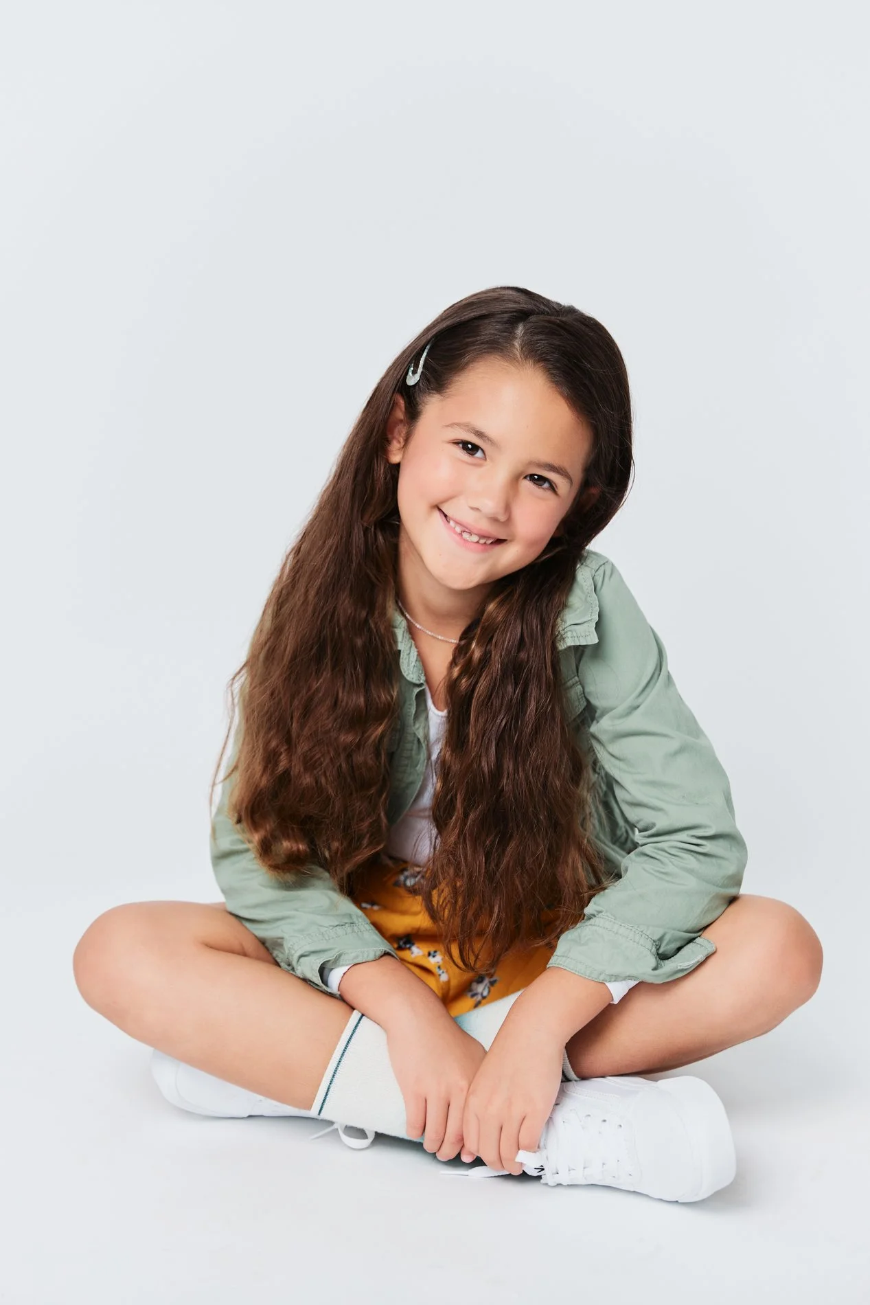Young girl with long brown hair, wearing a green shirt, yellow shorts, white socks, and white sneakers, sitting cross-legged against a plain light background, smiling.