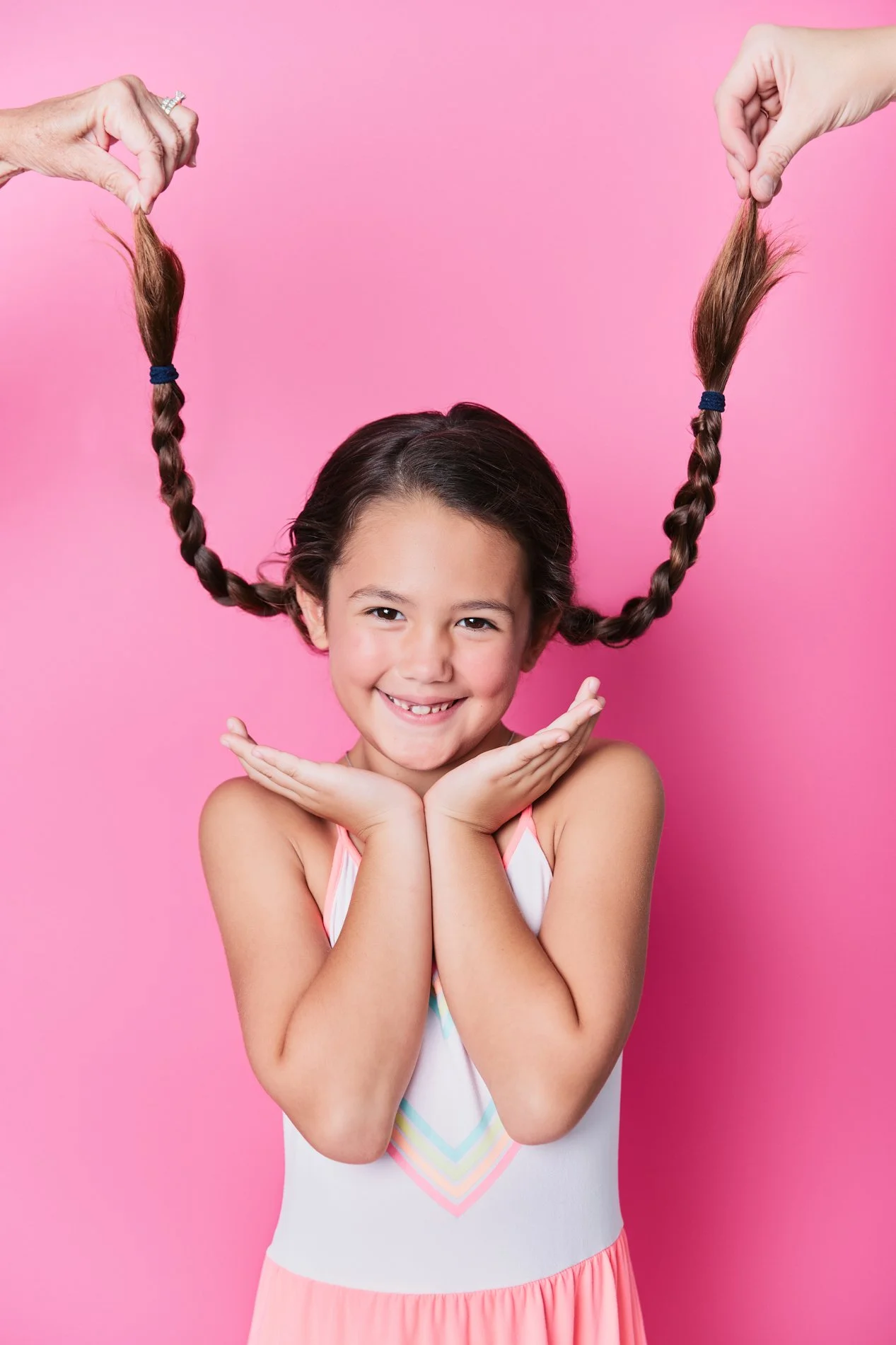 A young girl with dark hair smiling, holding her hands under her chin, with two hands holding her braided pigtails above her head against a pink background.