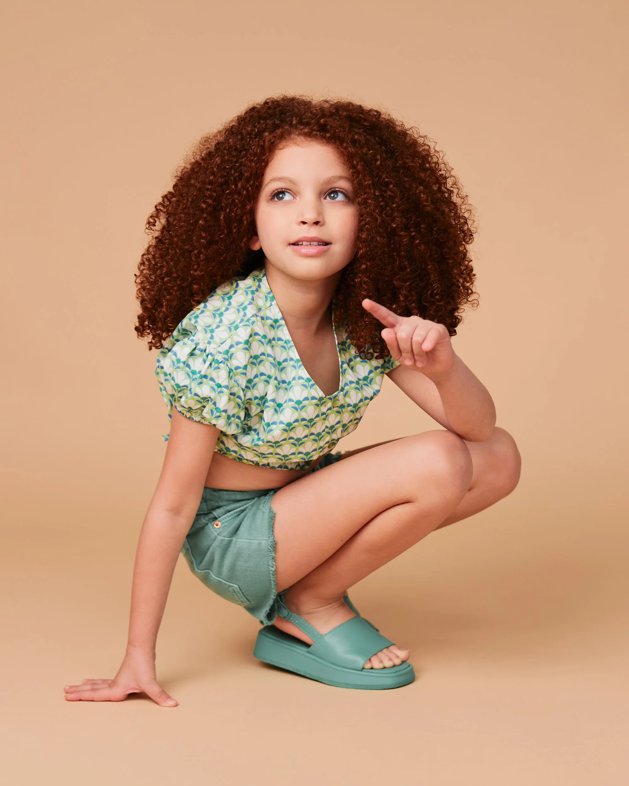 A young girl with curly red hair crouching with one hand on her knee and the other hand touching the ground, wearing a green patterned top, green shorts, and teal sandals, against a plain beige background.