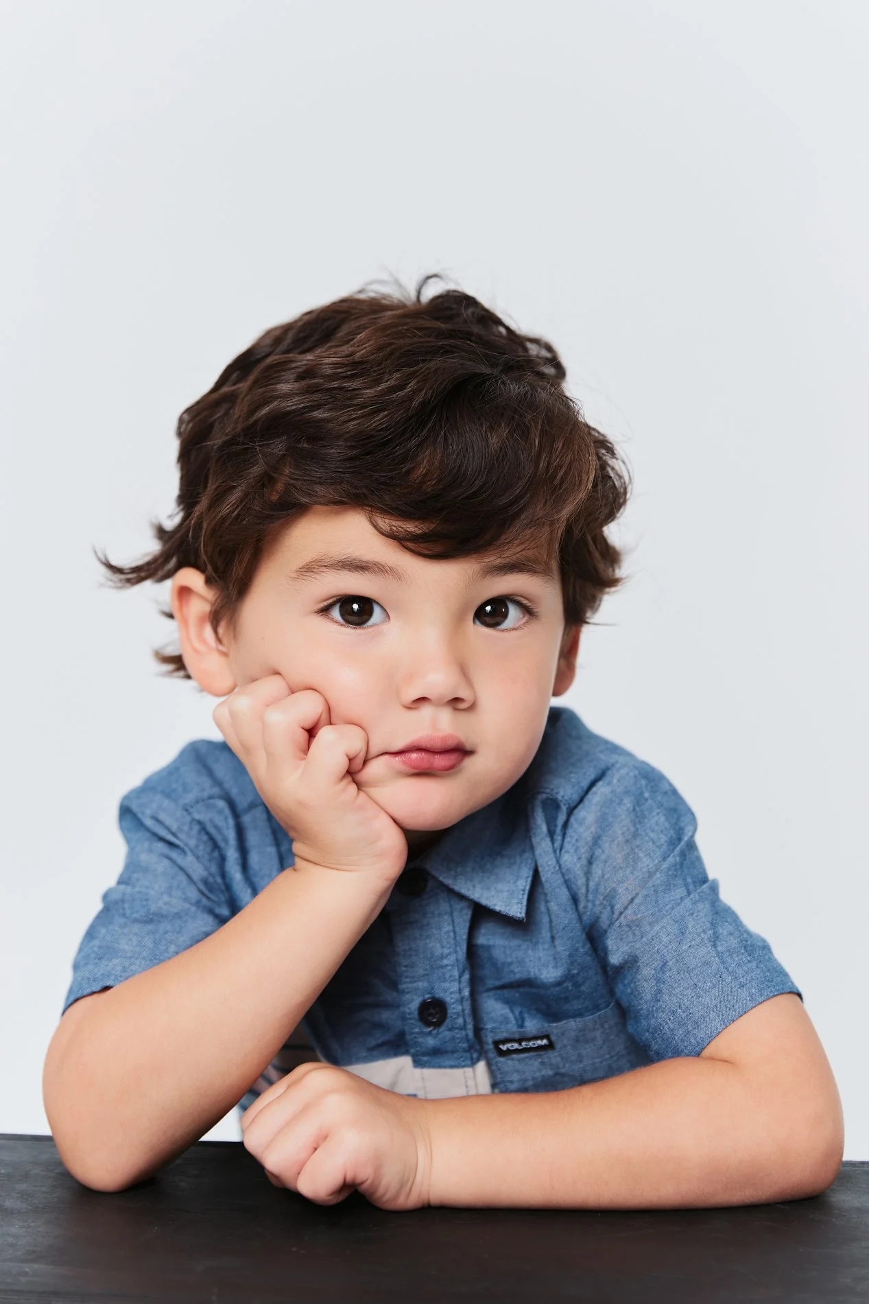 A young boy with dark brown hair and brown eyes, wearing a blue shirt, resting his chin on his hand while sitting at a black table against a plain white background.