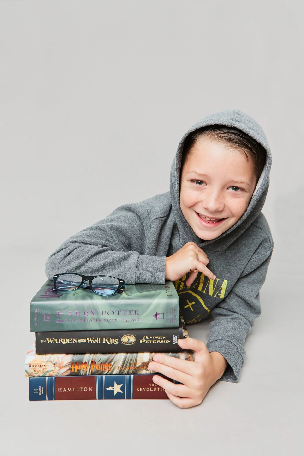 Young boy with a gray hoodie smiling and resting his head on a stack of four books, with glasses laid on top of the books, against a plain light gray background.