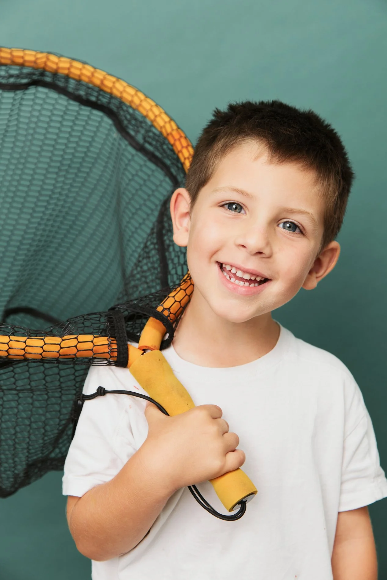 A smiling young boy with short dark hair and blue eyes holding a tennis racket over his shoulder against a teal background.