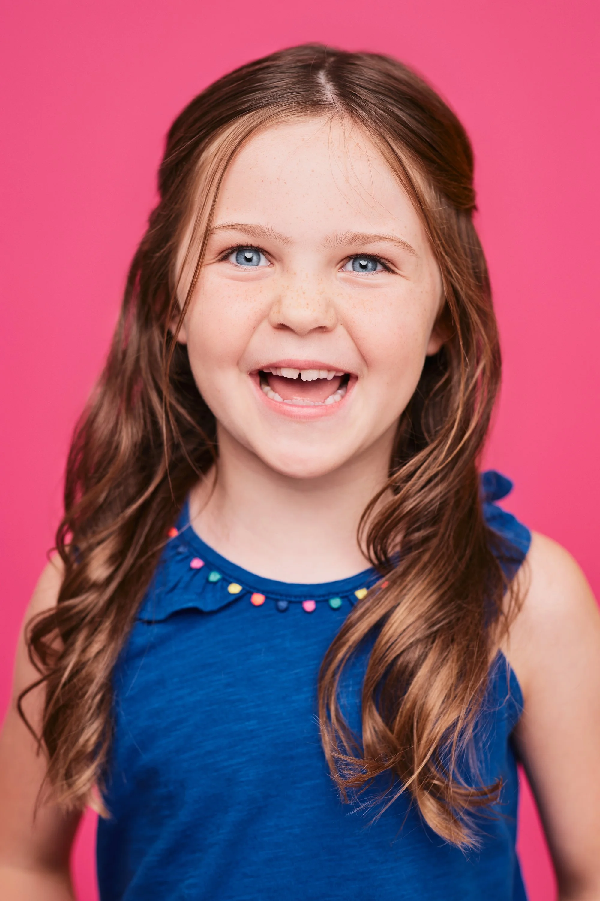 Smiling young girl with long wavy brown hair, blue eyes, freckles, wearing a blue sleeveless dress with colorful embellishments, against a bright pink background.