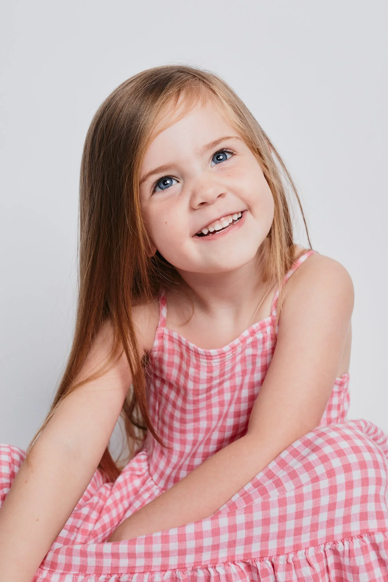 A young girl with red hair and blue eyes wearing a pink checkered dress, smiling and looking slightly upwards.