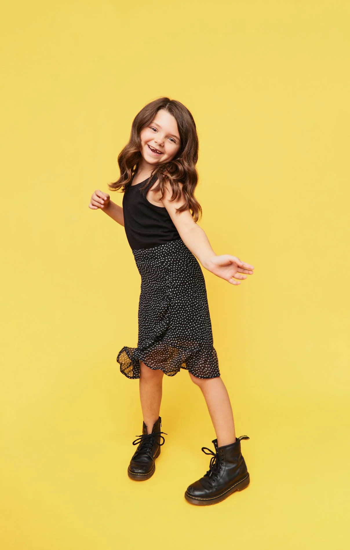 Houston School Photography | Young girl with brown hair smiling and dancing in front of a yellow background, wearing a black sleeveless top, polka-dotted skirt, and black boots.