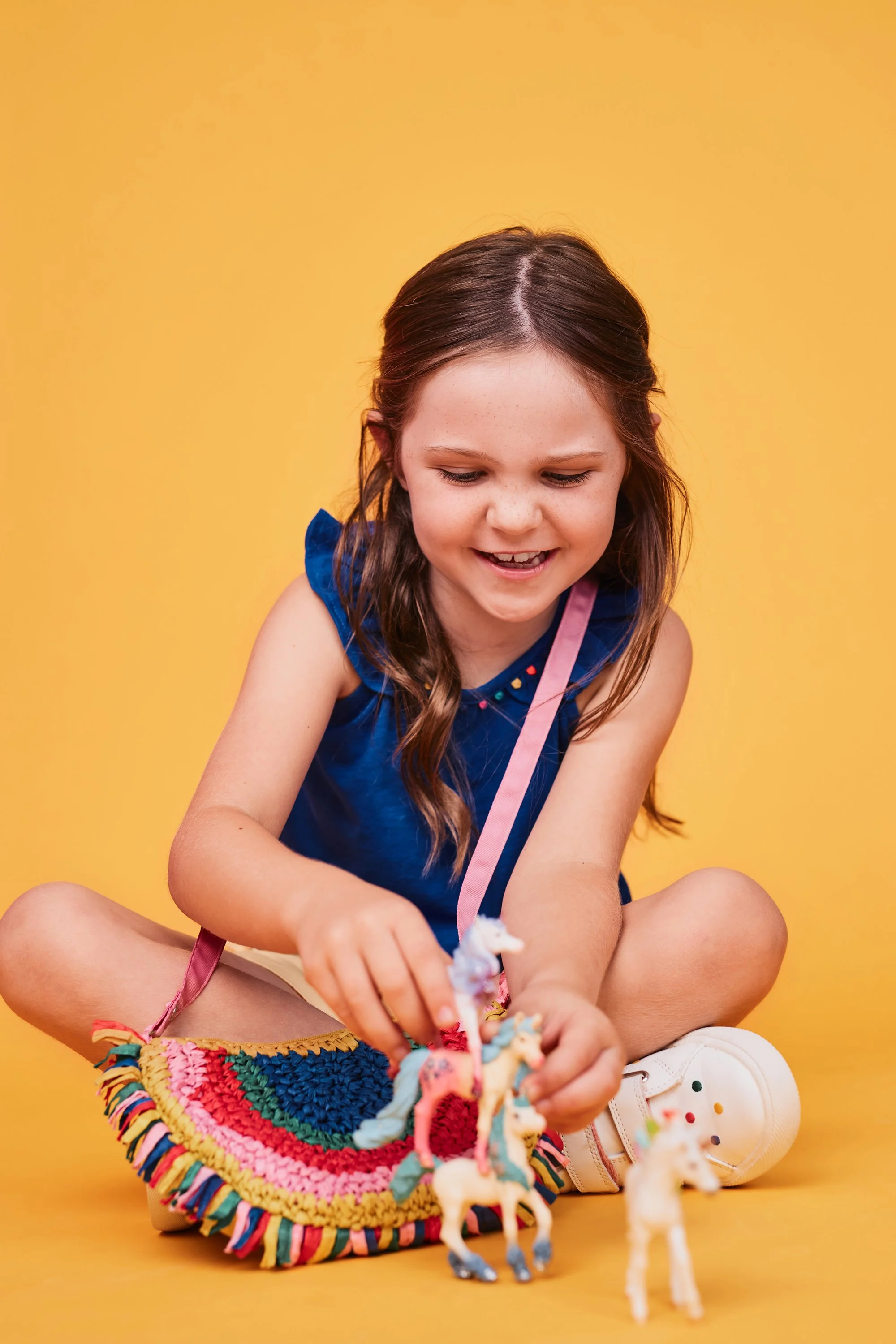 A young girl sitting cross-legged on the floor with a yellow background, playing with unicorn toys, wearing a blue top, white sneakers, and rainbow-colored skirt.
