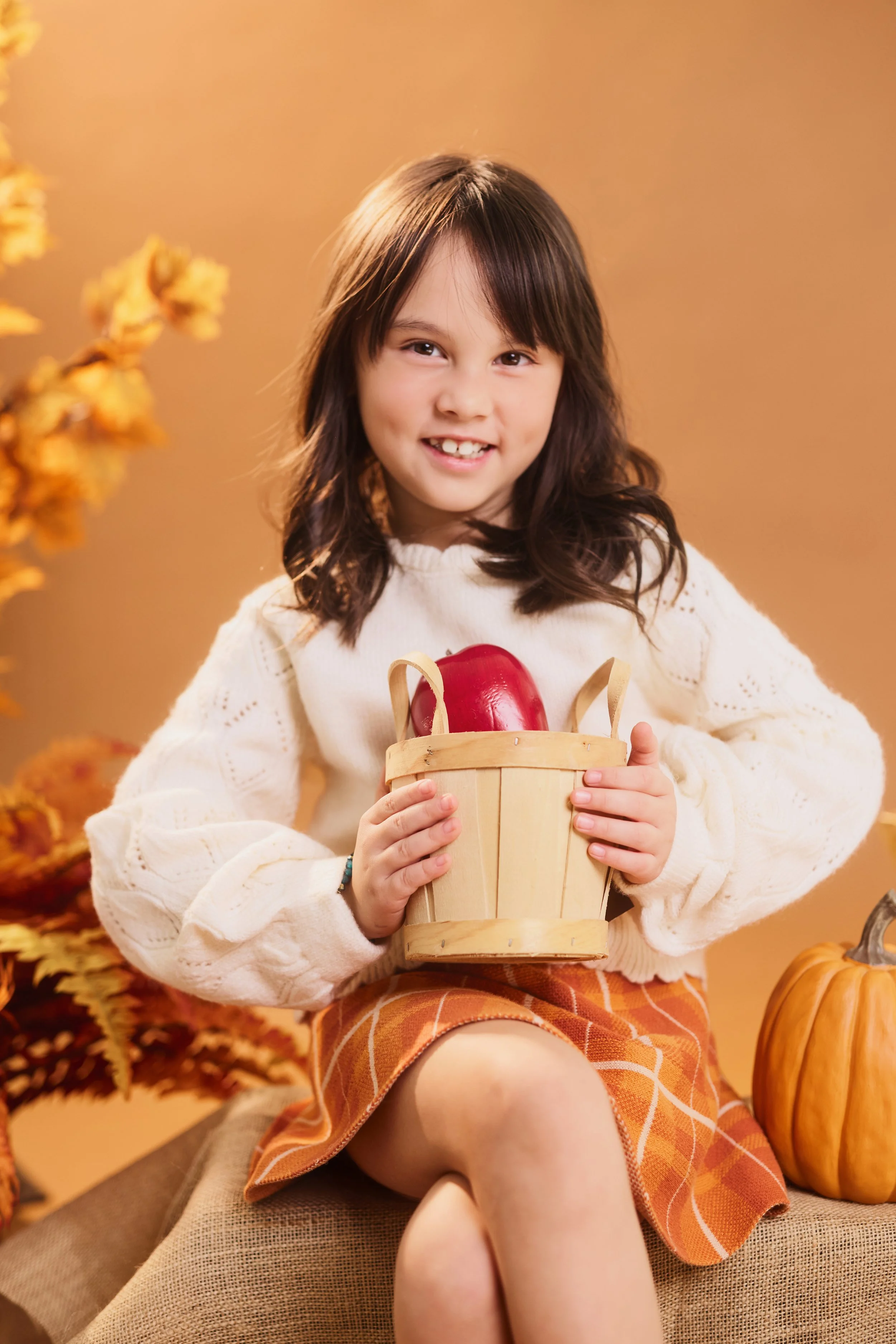 Houston School Photography for Preschool | A young girl with brown hair and a white sweater holding a small wooden basket with a red apple inside. She is sitting in a fall-themed setting with orange and yellow leaves, pumpkins, and a warm background.