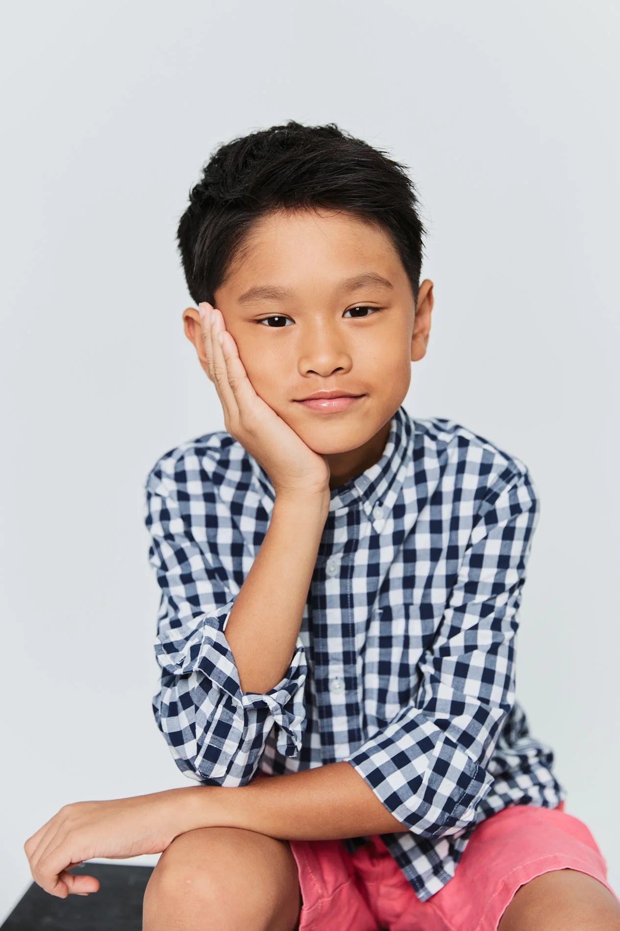 Houston Children's Photographer | A young boy with dark hair and tan skin, wearing a blue and white checkered shirt and pink shorts, sitting with his right arm resting on his knee and his left hand supporting his face