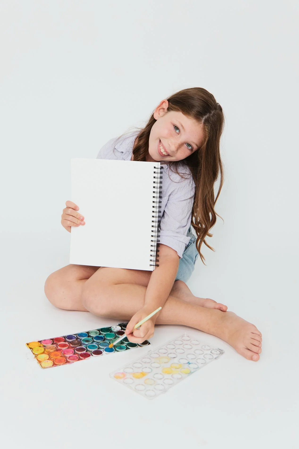 A young girl sitting cross-legged on the floor, holding a blank spiral notebook and a paintbrush, with watercolor paints and palettes spread out in front of her, engaging in painting.