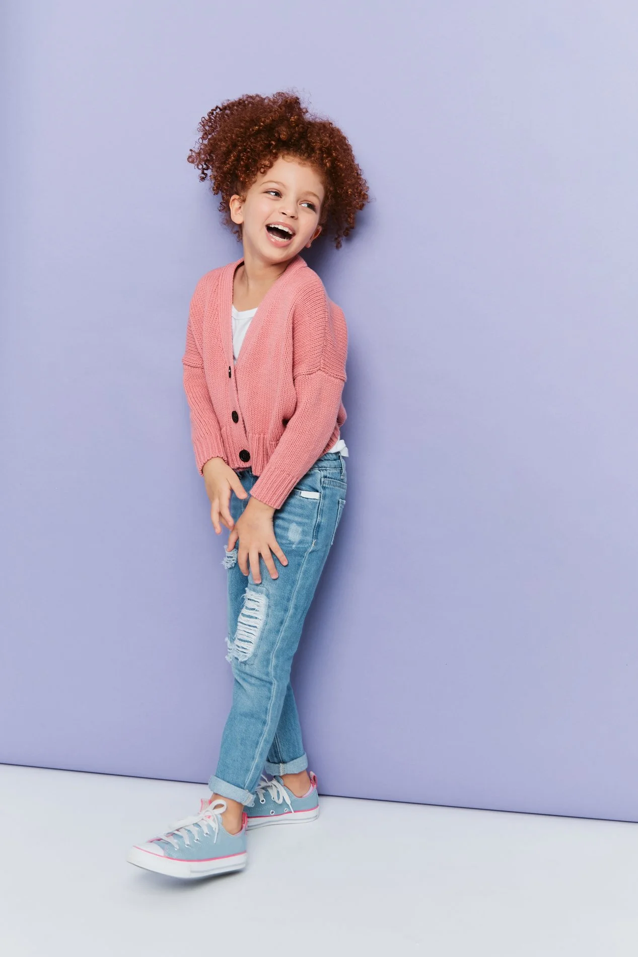 Houston School Photographer young girl with curly red hair wearing a pink cardigan, white T-shirt, ripped jeans, and sneakers, laughing against a purple background.