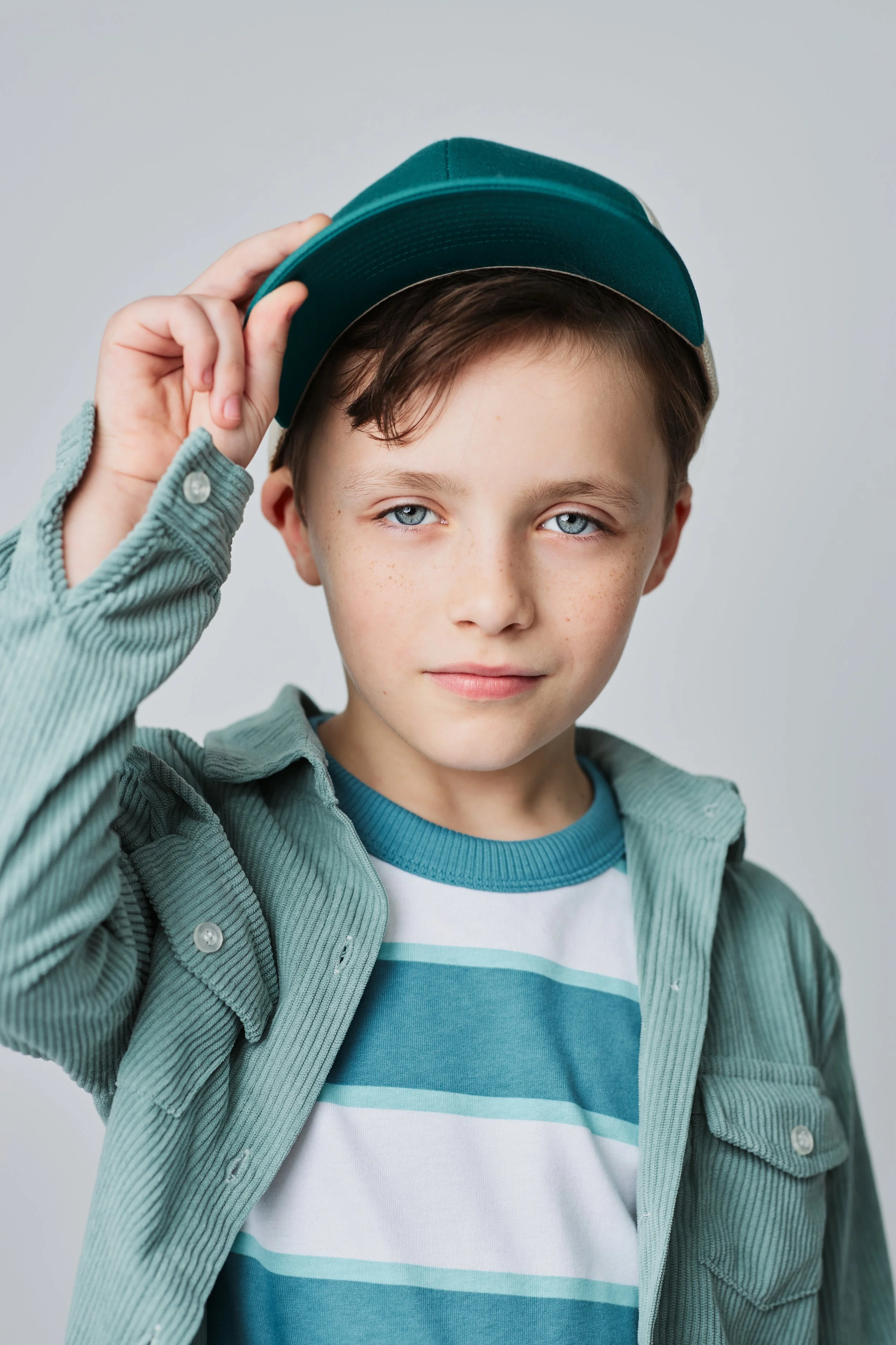 Houston Children's Photographer | A young boy with light brown hair and blue eyes adjusting a teal baseball cap, wearing a teal and white striped t-shirt with a greenish-gray jacket over it, against a plain light gray background.