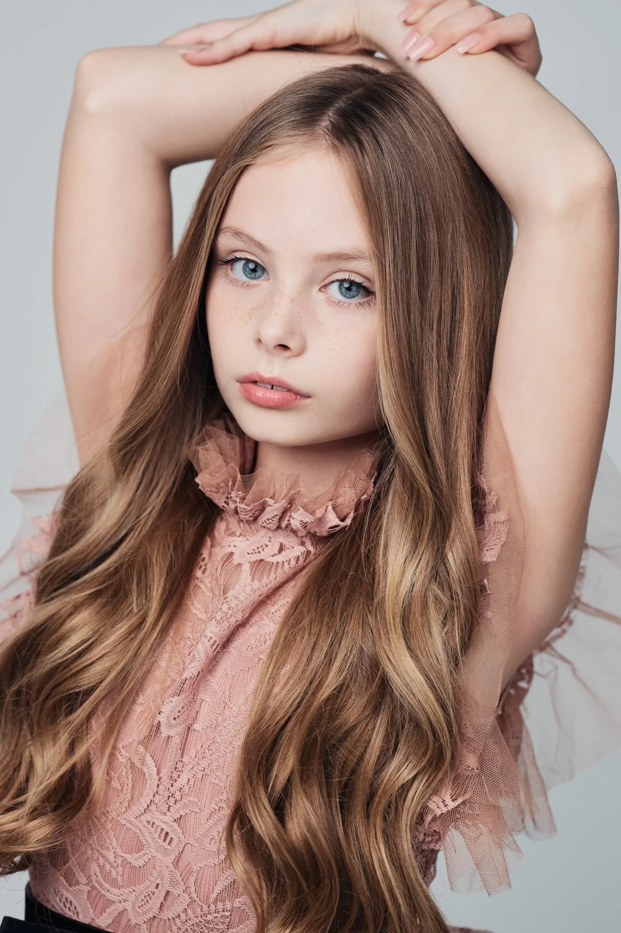 A young woman with long, wavy brown hair and blue eyes poses with her arms raised above her head, wearing a pink lace top with ruffled details around the neckline and sleeves.