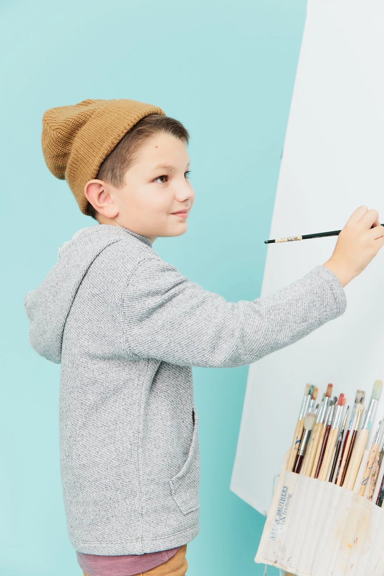 A young boy wearing a brown knit beanie and a gray hoodie paints on a whiteboard. There are paintbrushes stored in a container to the right.