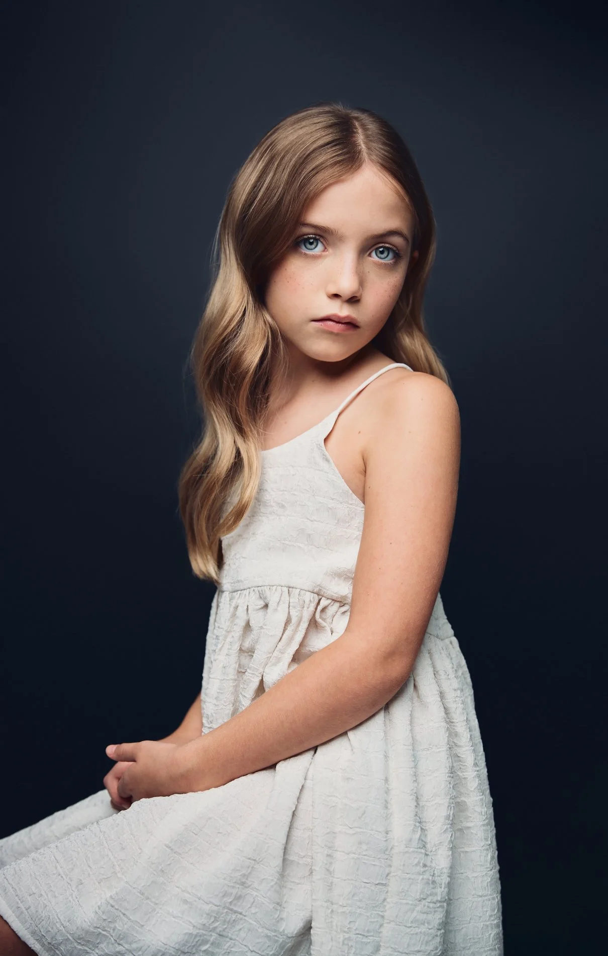 Young girl with long light brown hair and blue eyes, wearing a white dress, sitting against a dark background.