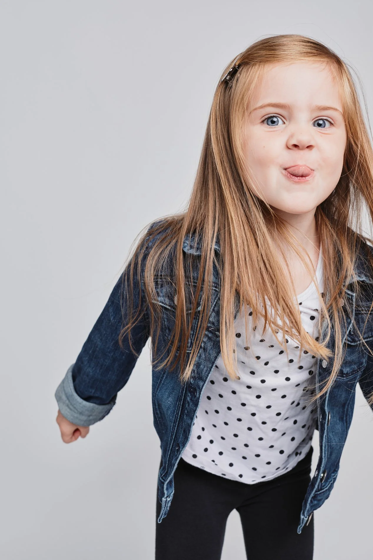 A young girl with long red hair, blue eyes, and fair skin sticking out her tongue, wearing a denim jacket over a white polka dot shirt and black pants, standing against a plain gray background.