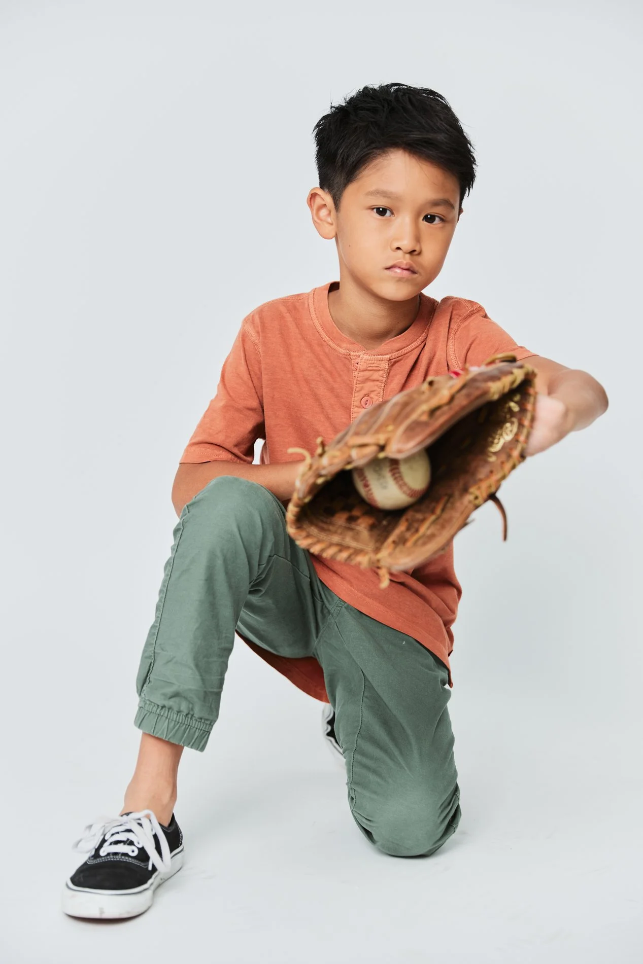 Young boy in orange shirt and green pants, wearing sneakers, kneeling with one knee on the ground, holding a baseball glove with a baseball inside, against a plain light background.
