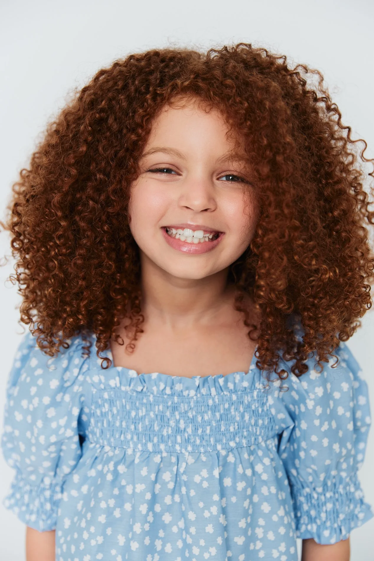 Smiling young girl with curly red hair wearing a blue dress with a white floral pattern.