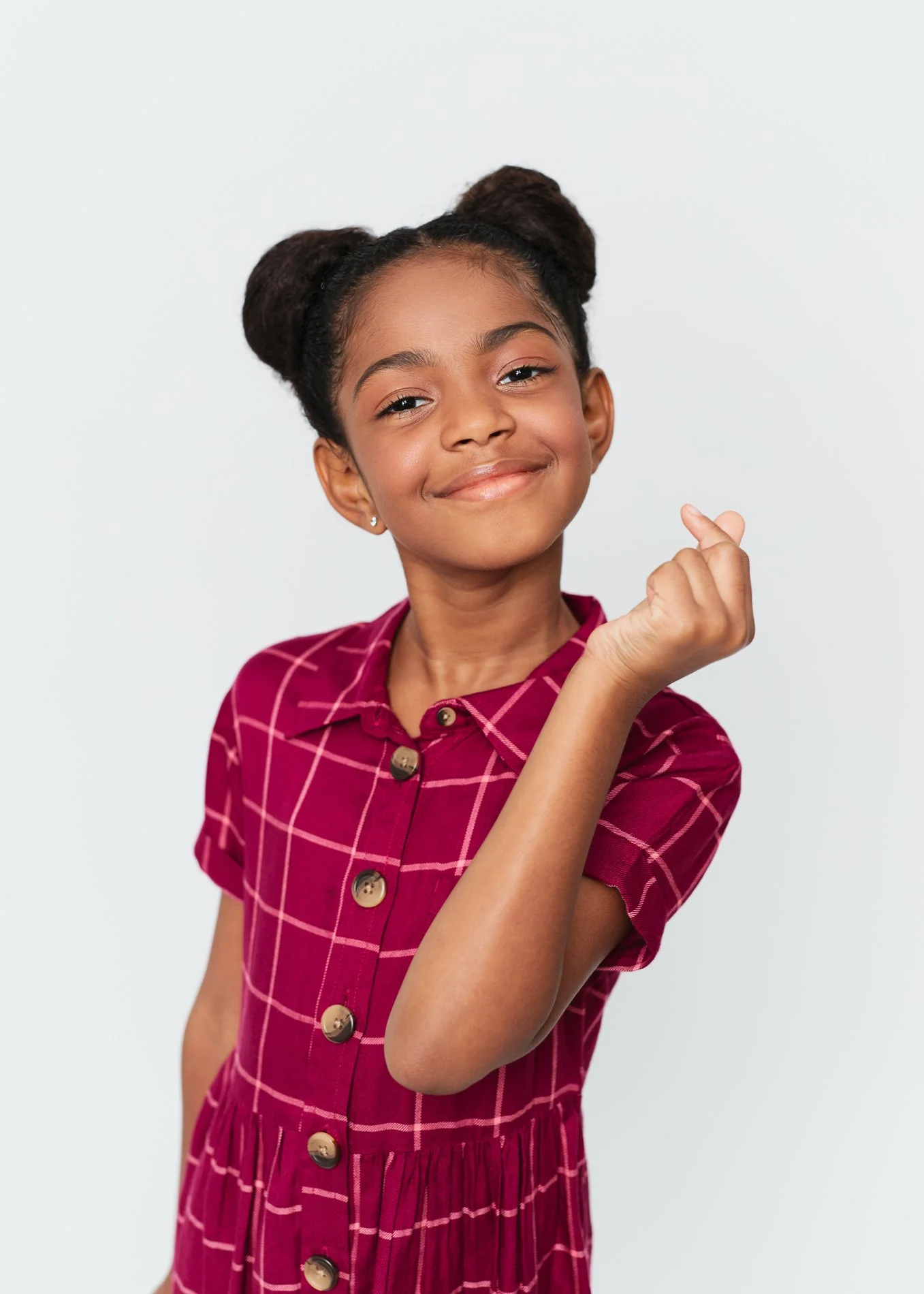 A young girl with two puffs in her hair, wearing a maroon dress with a checkered pattern, smiling and making a finger heart gesture against a plain white background.