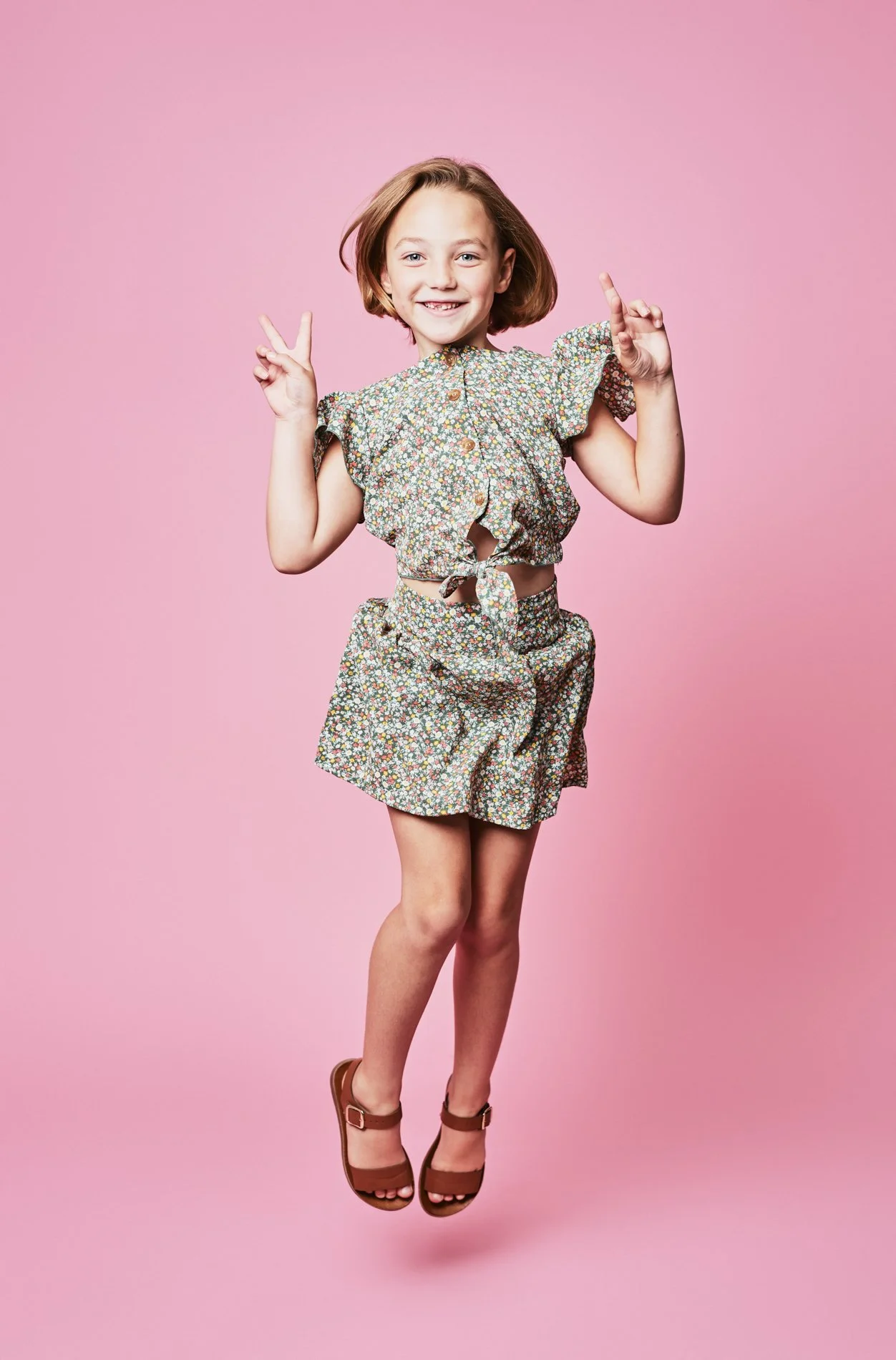 Young girl smiling, making peace signs with both hands, wearing a floral crop top and skirt, brown sandals, against a pink background.