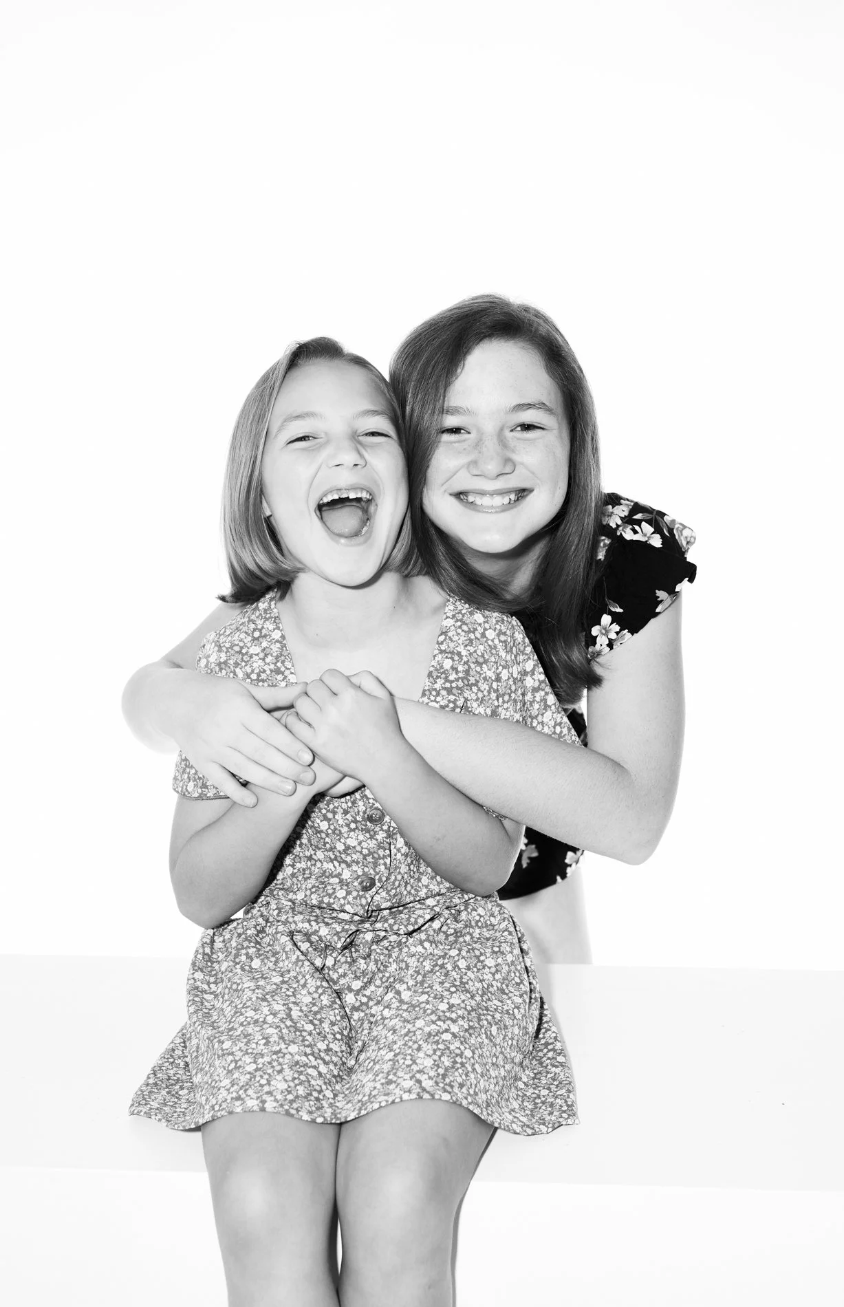 Two young girls, one with short hair and the other with long hair, smiling and hugging each other, sitting against a plain white background.