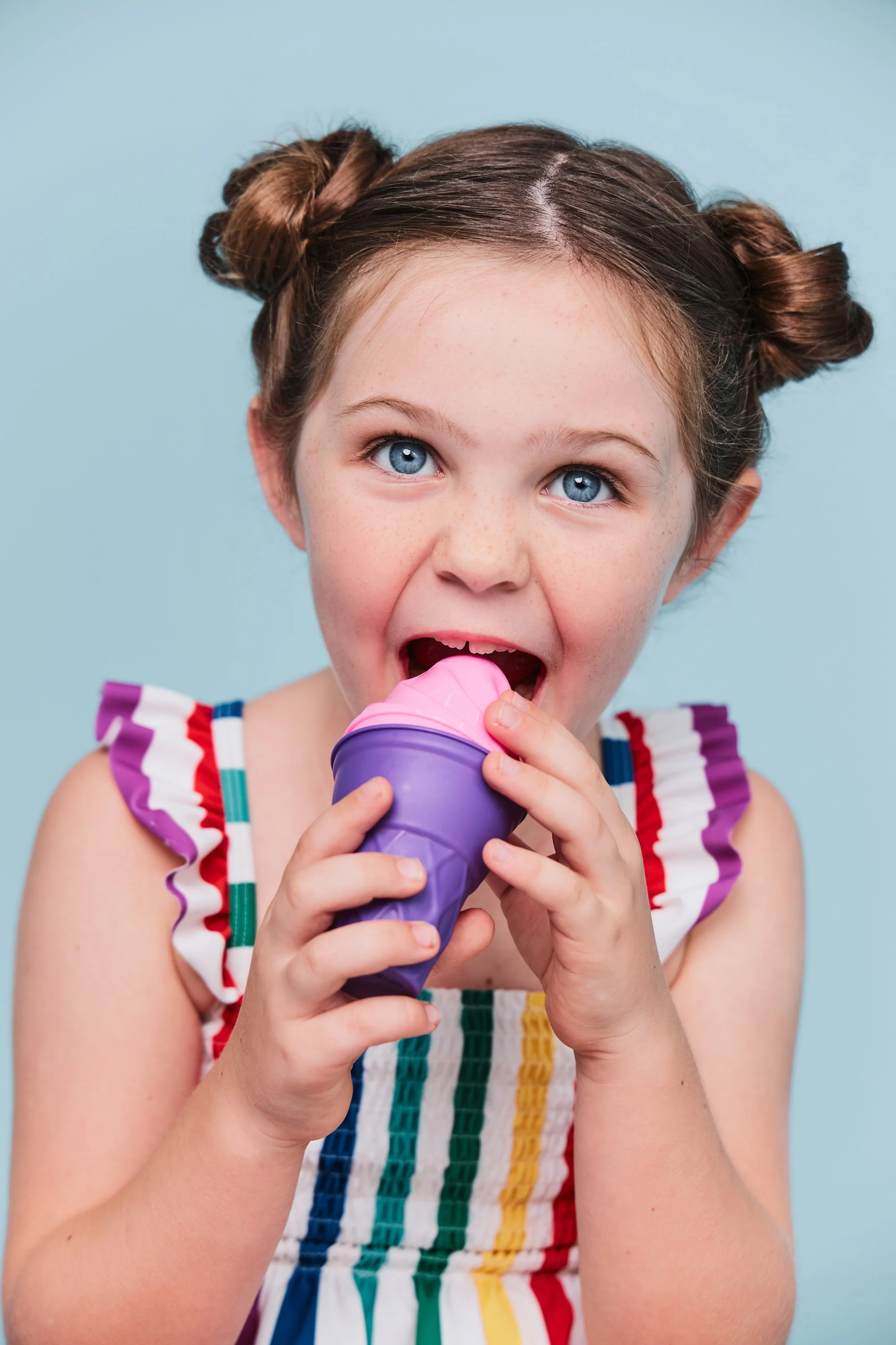 A young girl with blue eyes and brown hair styled in two buns, wearing a colorful striped dress, is eating ice cream from a purple cone against a light blue background.