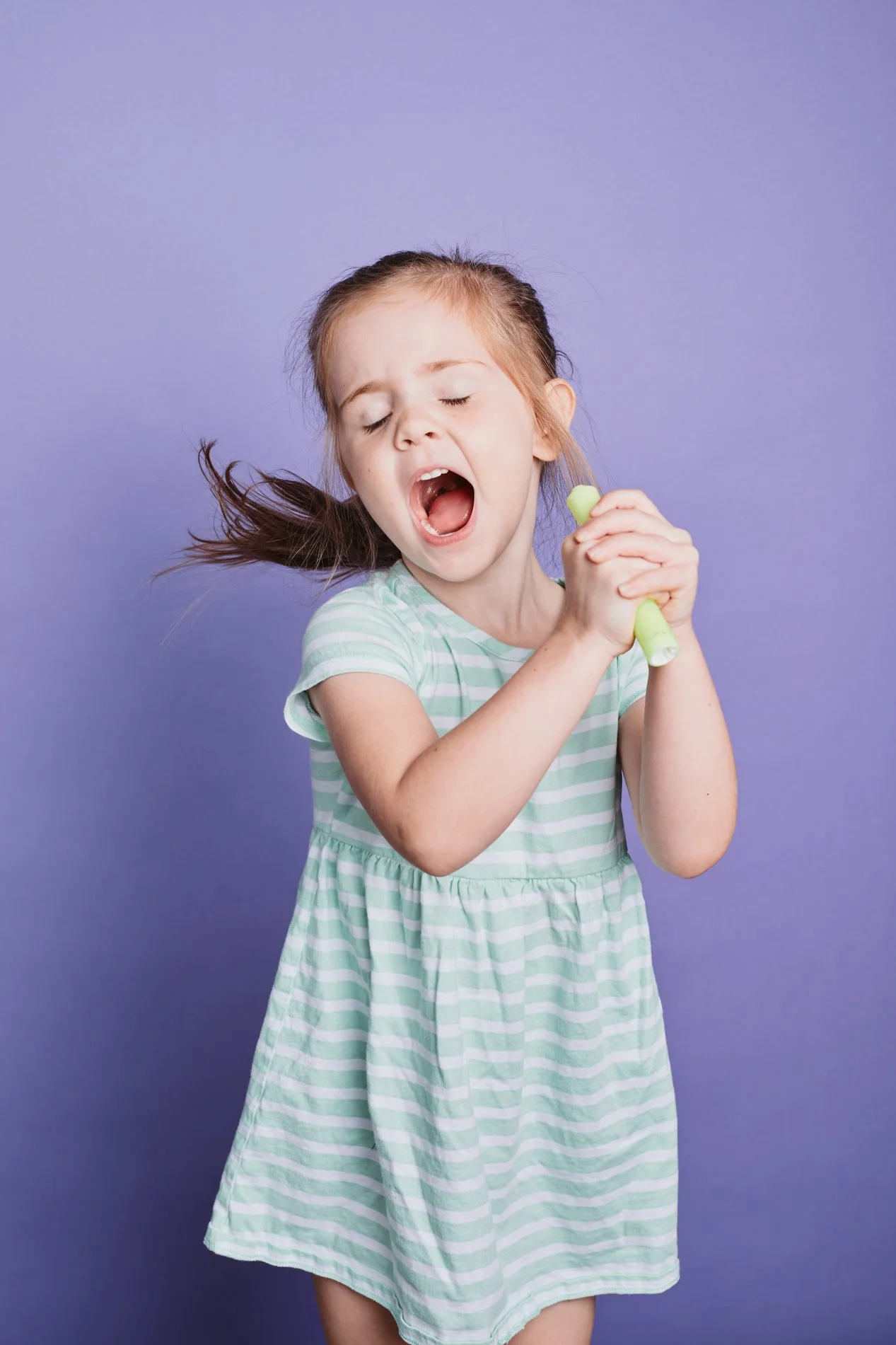 Young girl with closed eyes and open mouth, holding a green object, standing against a purple background.
