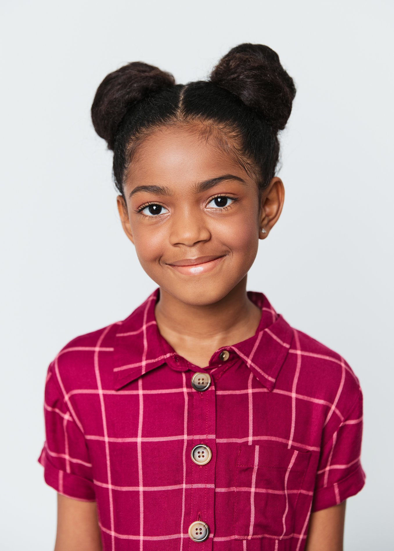 A young girl with dark hair styled in two buns, wearing a red plaid shirt with beige buttons, smiling softly against a plain, light-colored background.