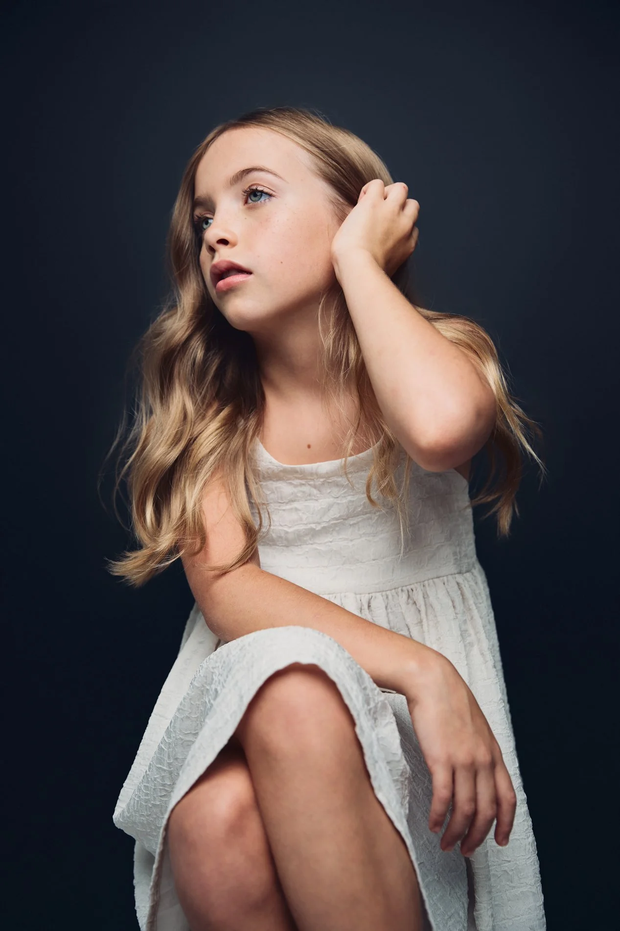 A young woman with long, wavy blonde hair and blue eyes sits against a dark background, wearing a white textured dress. She rests her hand on her head and looks thoughtfully into the distance.