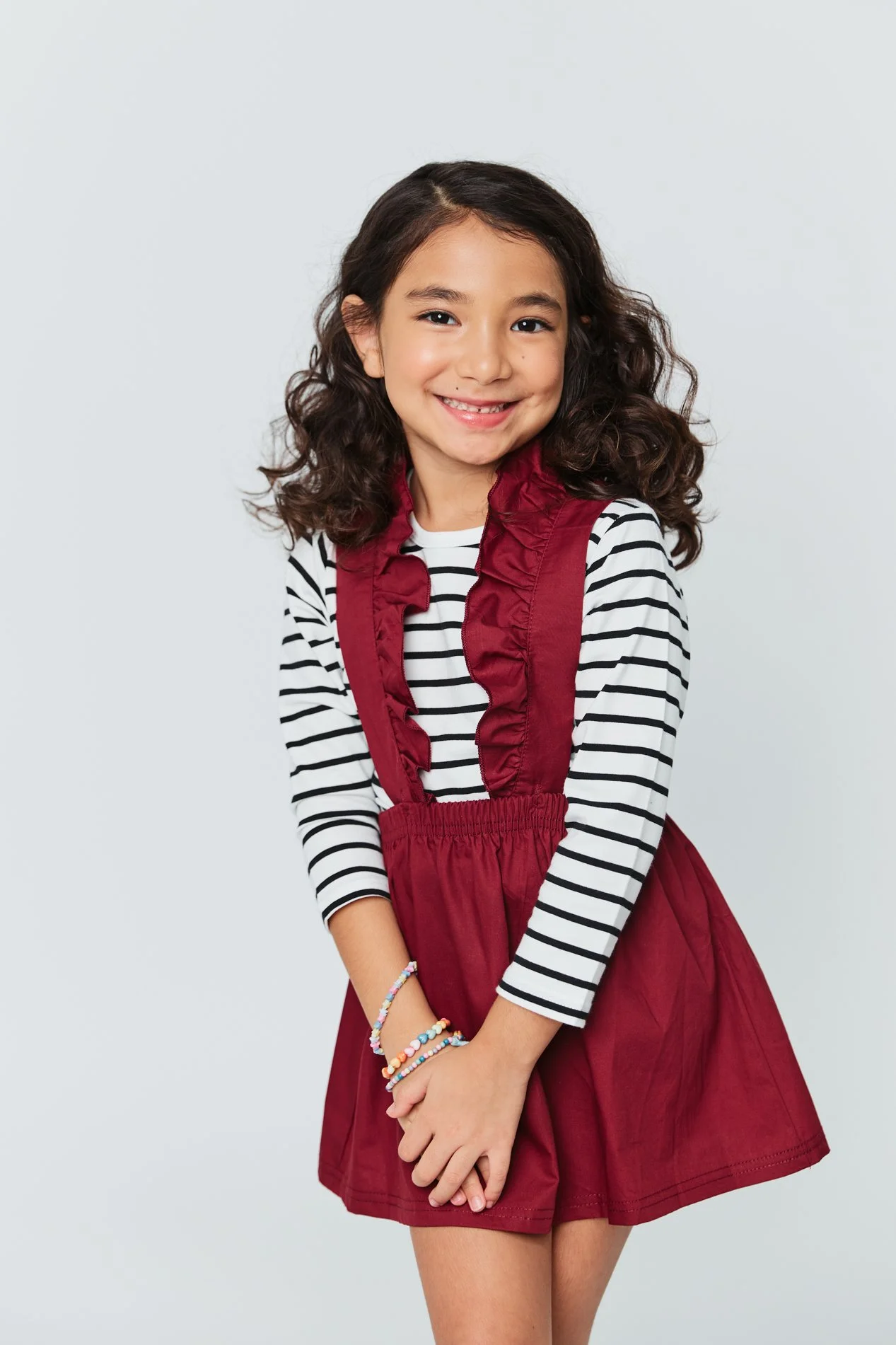 A young girl with curly dark hair, smiling, wearing a striped long sleeve shirt, a red dress, and colorful beaded bracelets against a plain light background.