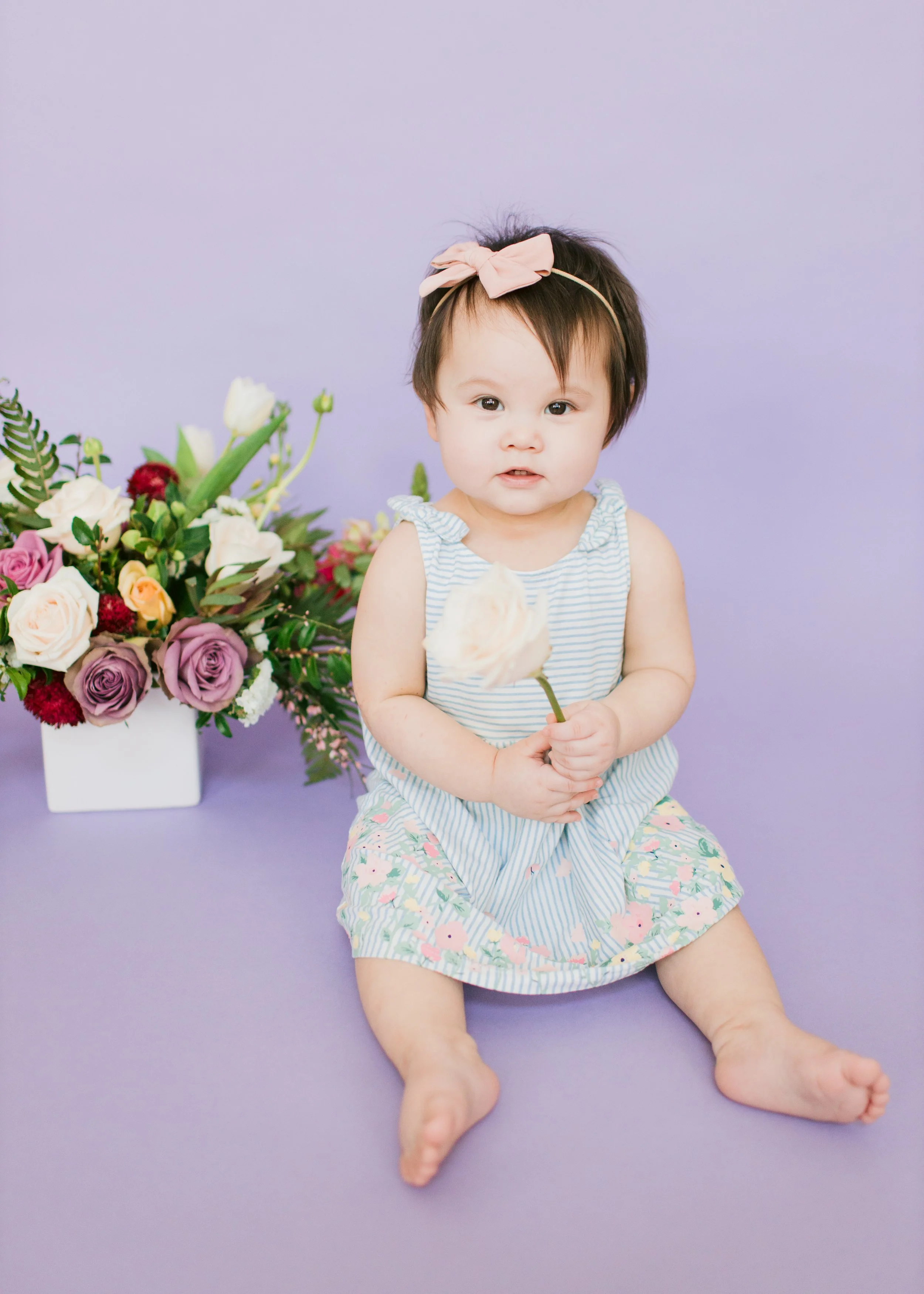A young child sitting on a purple surface, holding a white flower, with a large bouquet of various colored flowers to her left, wearing a striped dress with ruffled sleeves and floral shorts, and a pink bow headband, against a light purple background
